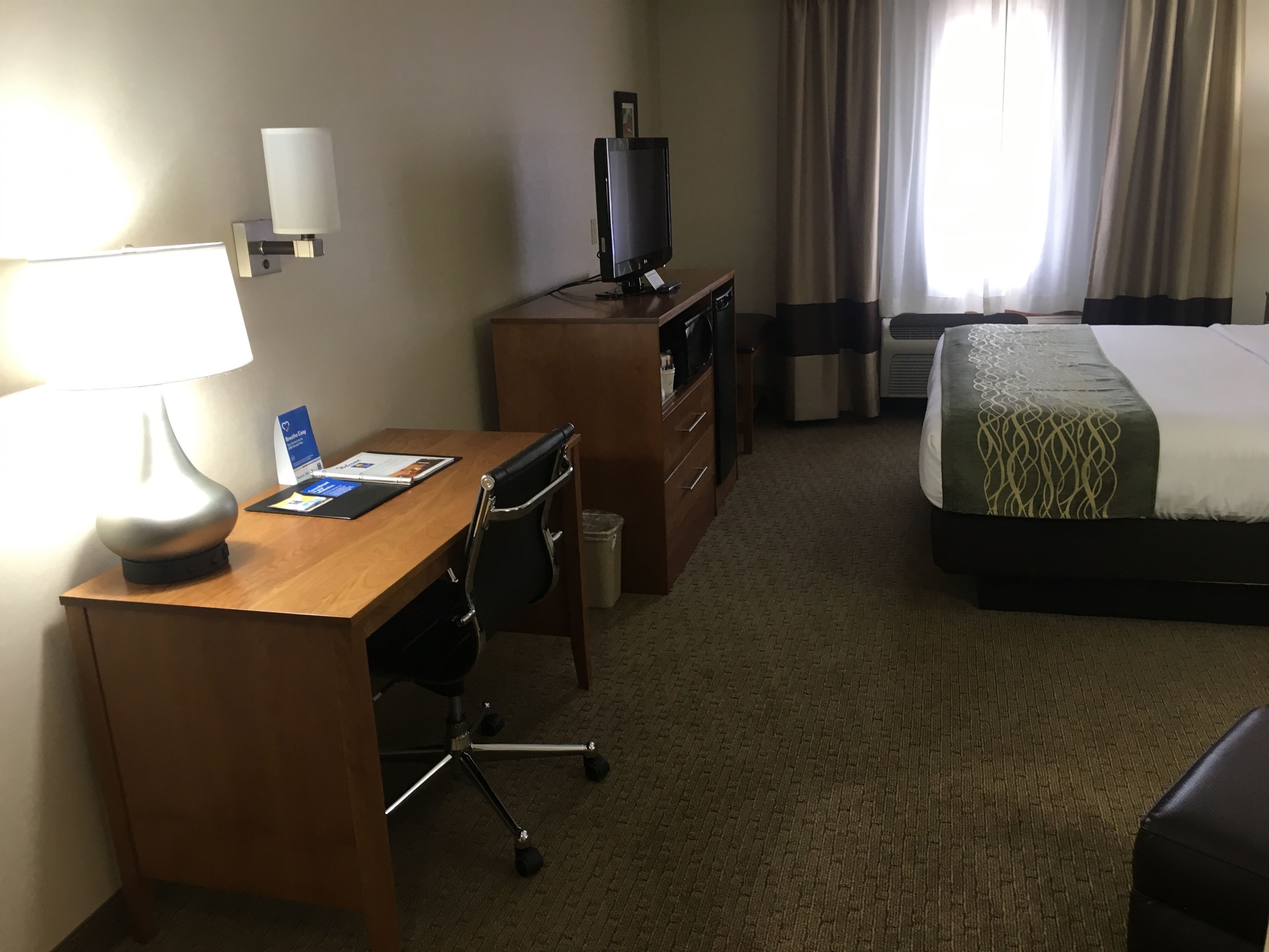 A hotel room with a wooden desk, a chair, a television, a bed, and a window with curtains.