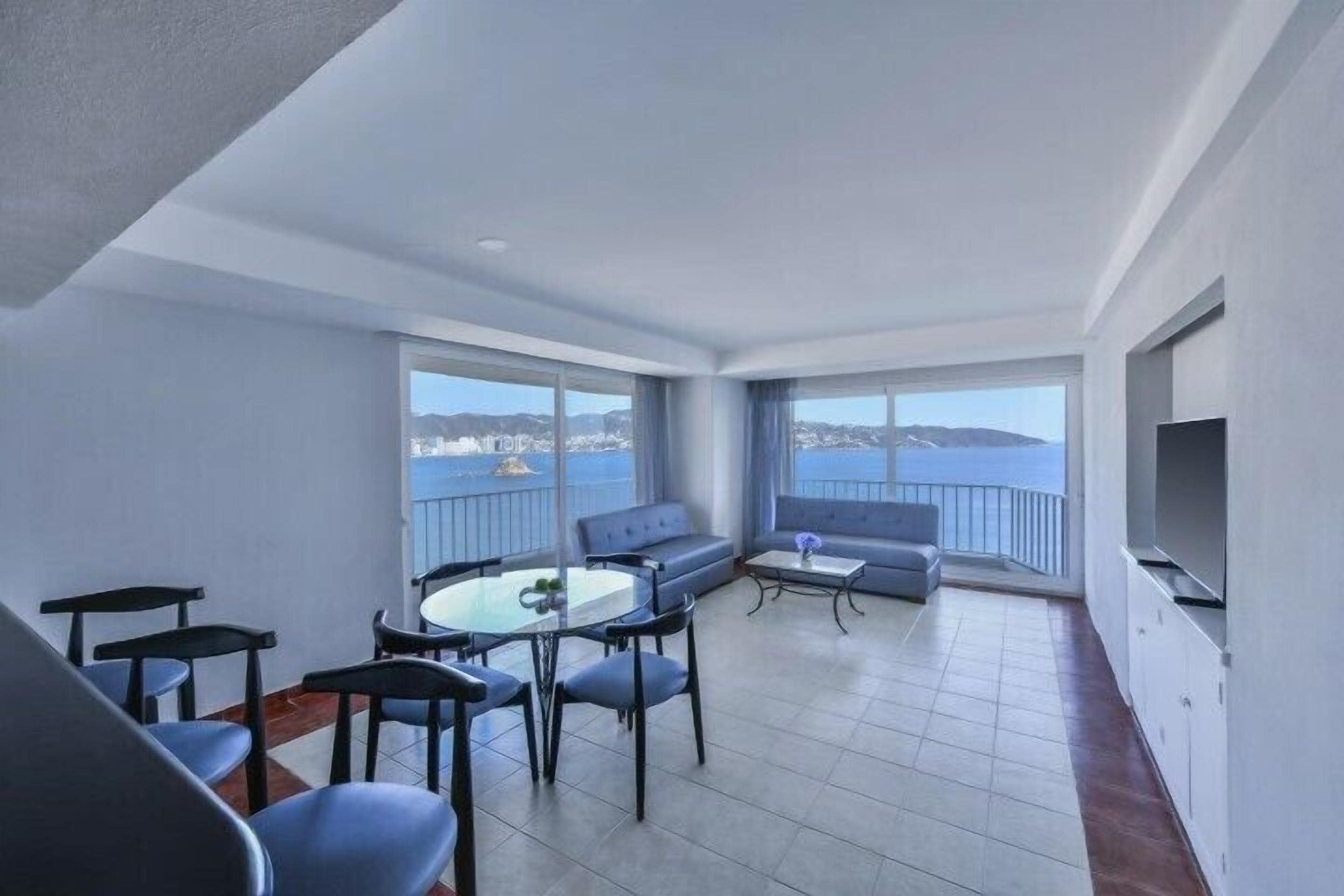 A balcony with a view of the sea, a seating area with a table and chairs, and a flat-screen TV mounted on the wall.