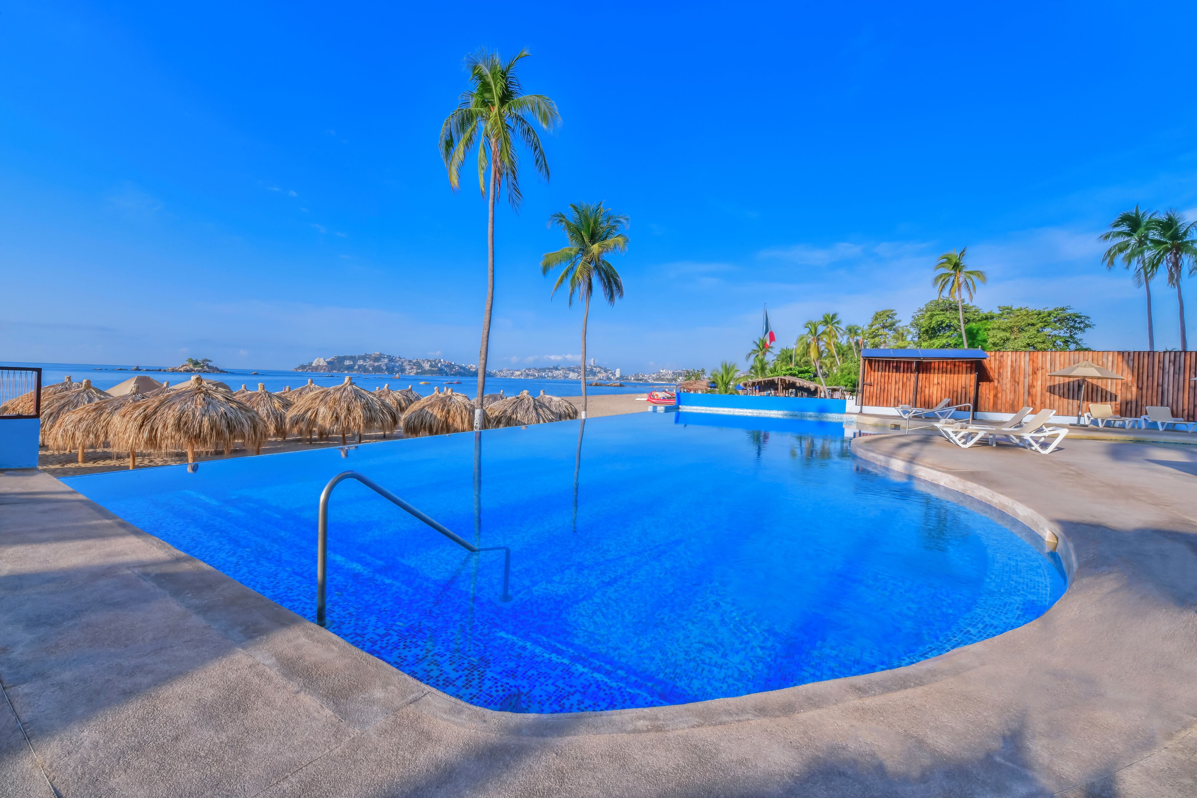 Una piscina con agua azul cristalina, rodeada de sillas de descanso y palmeras, con vista al océano y un horizonte urbano lejano.