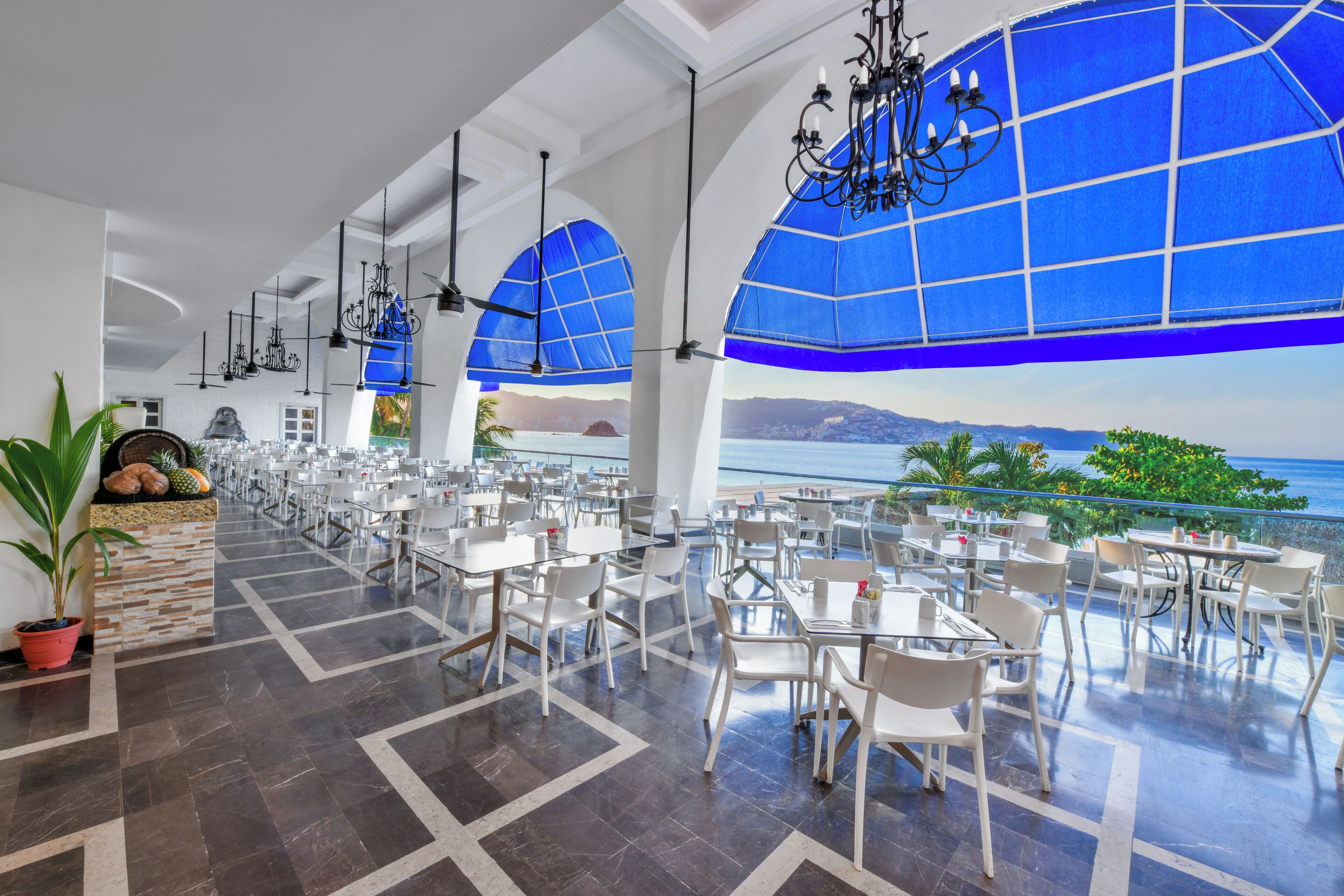 Un comedor espacioso con mesas y sillas blancas, amplias ventanas azules y vista al mar.