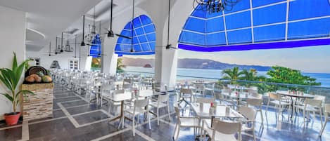 A spacious dining area with white tables and chairs, large blue windows, and a view of the sea.