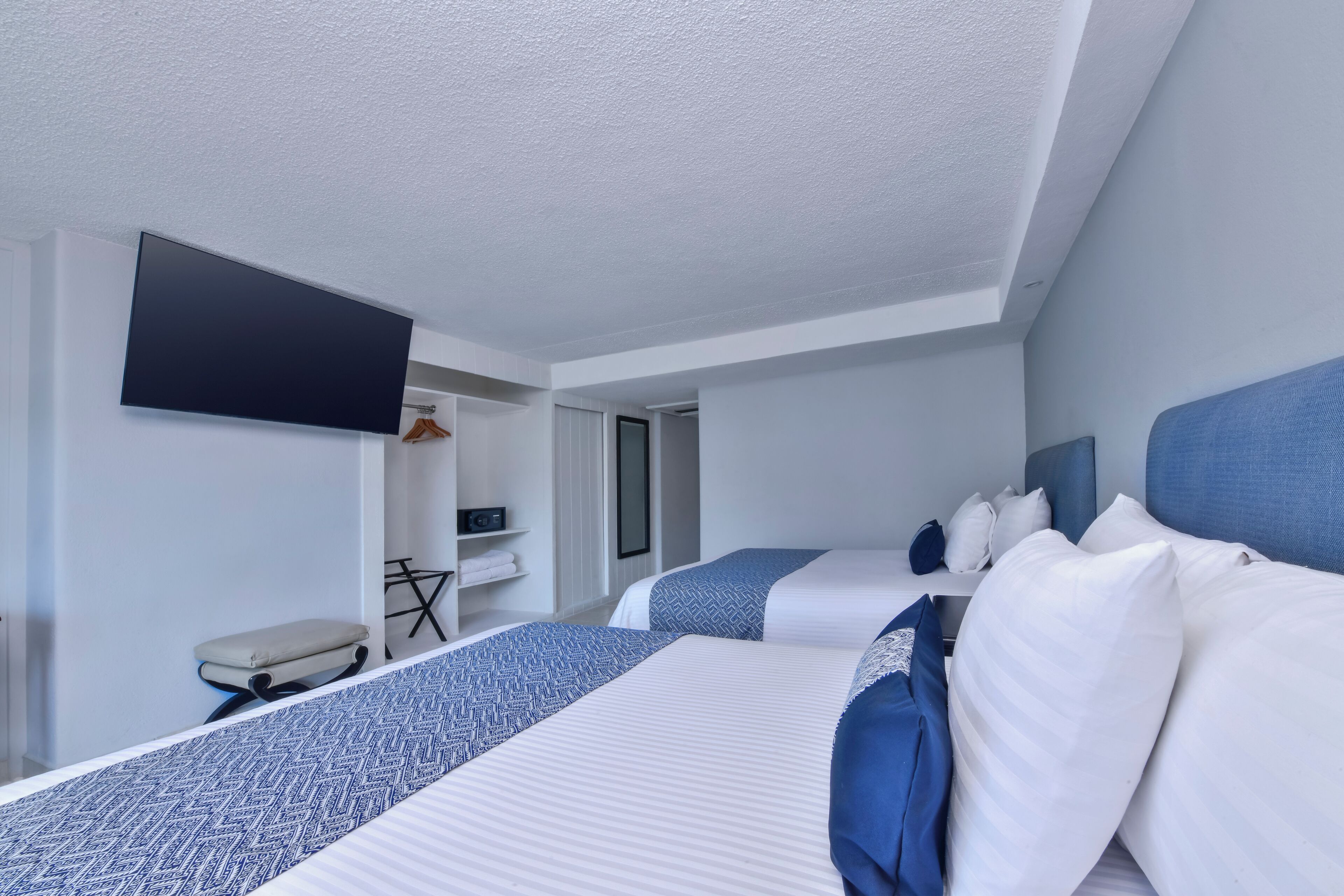 Habitación de hotel con dos camas, un televisor de pantalla plana montado en la pared, un escritorio pequeño con una silla y un armario.