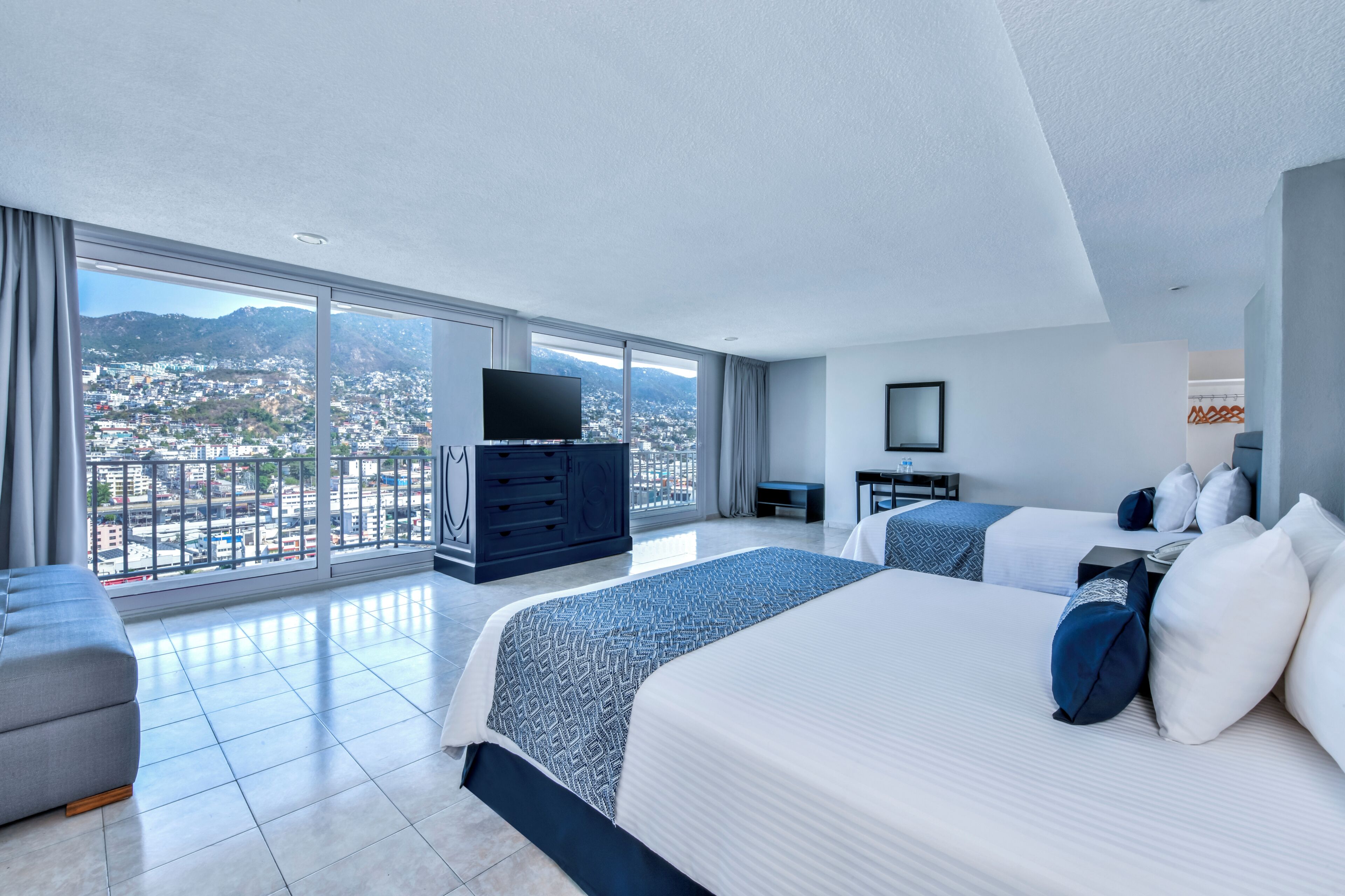 A modern hotel room with a large bed, a TV, a desk, and a view of a cityscape.