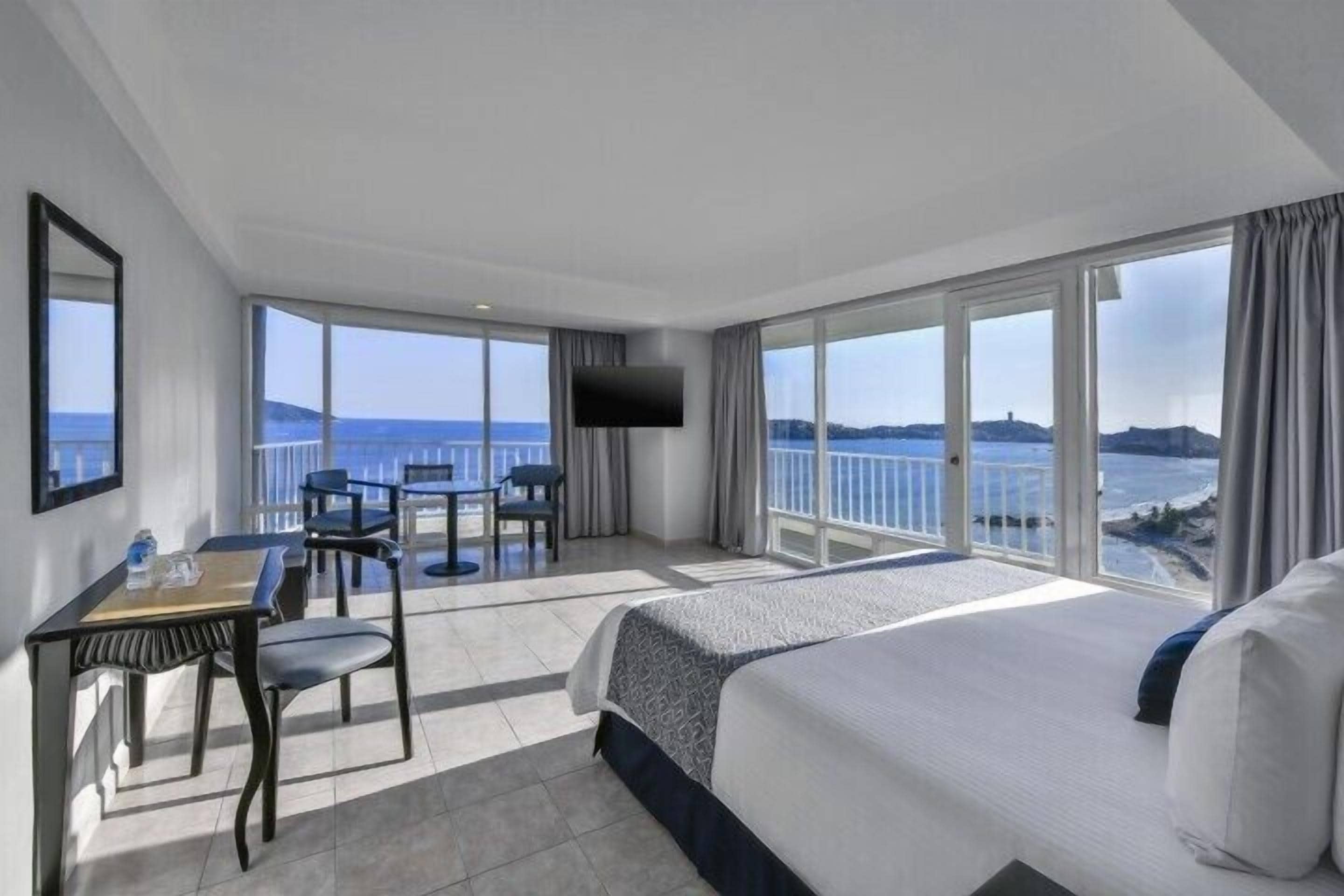Une chambre d’hôtel équipée d’un lit, d’un bureau, d’une chaise, d’une télévision et de grandes fenêtres donnant sur la mer.