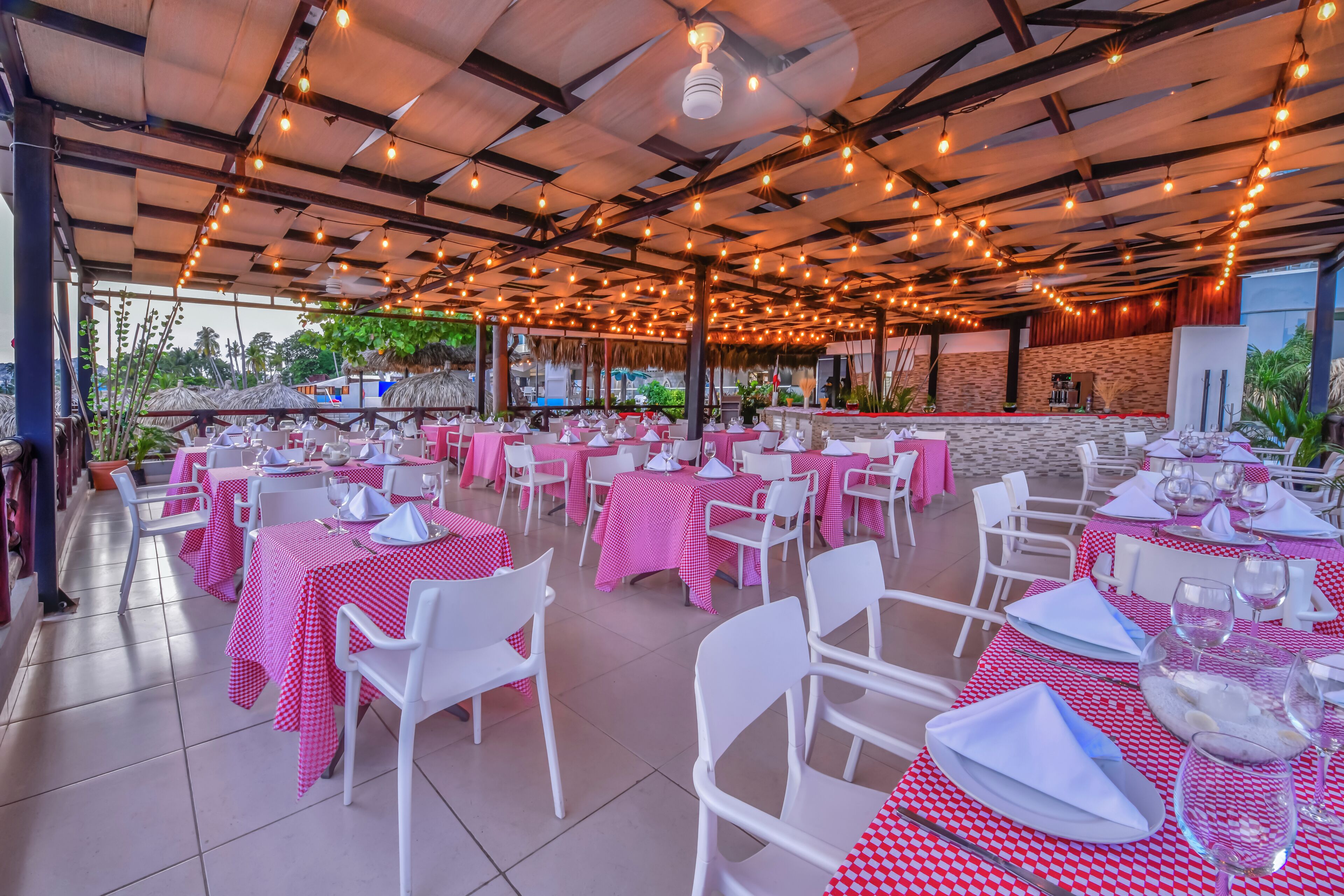 Un área de comedor al aire libre con mesas cubiertas con mantel de cuadros rojos y blancos, sillas blancas y luces colgantes.