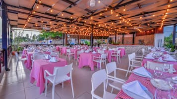 Un área de comedor al aire libre con mesas cubiertas con mantel de cuadros rojos y blancos, sillas blancas y luces colgantes.