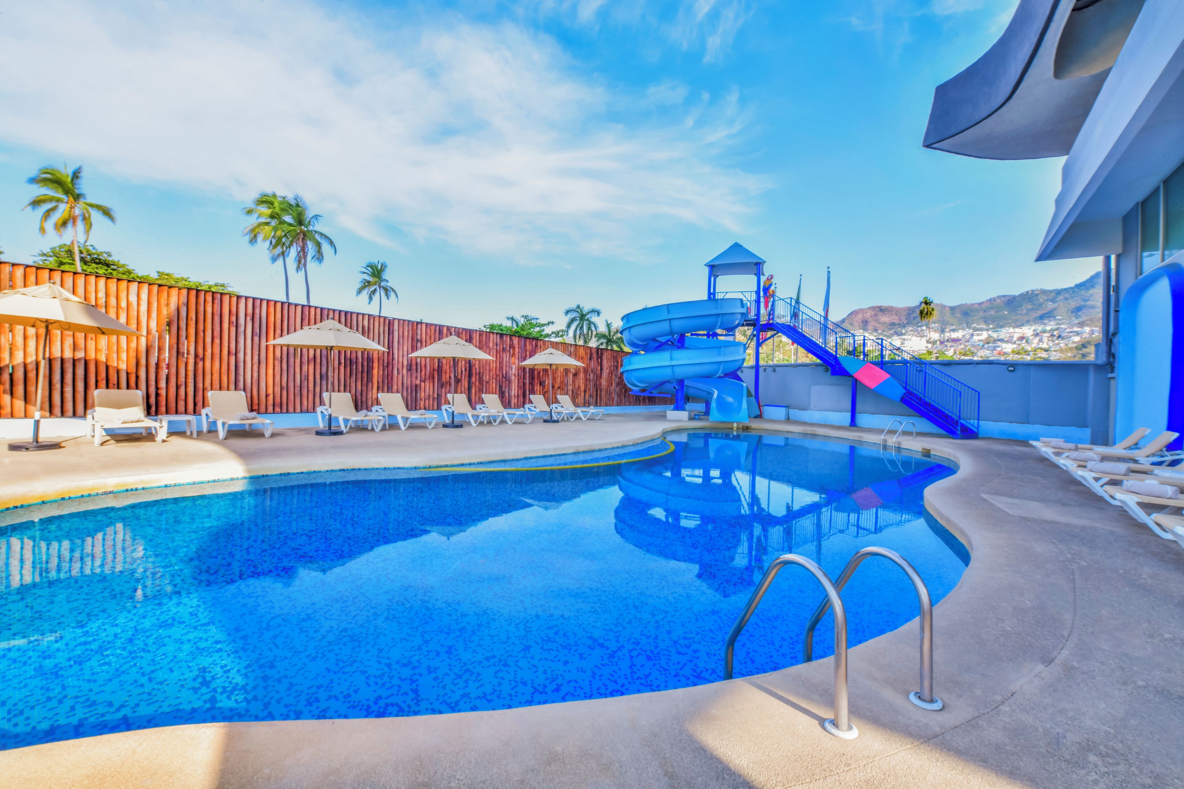A swimming pool with a water slide, lounge chairs, umbrellas, and a view of palm trees and mountains.