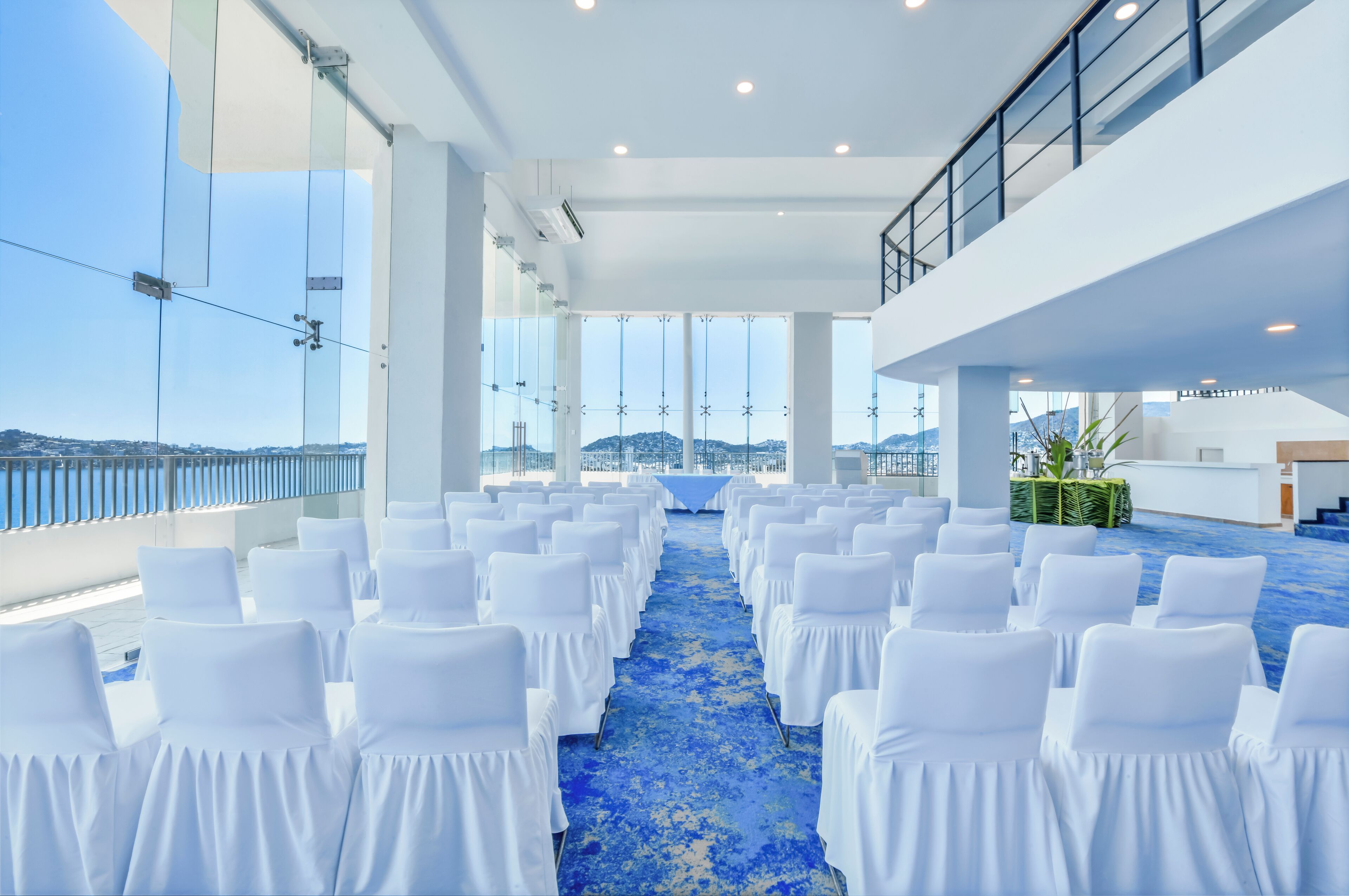 Une salle de conférence avec des rangées de chaises blanches, une moquette bleue, de grandes fenêtres et une vue sur les montagnes.