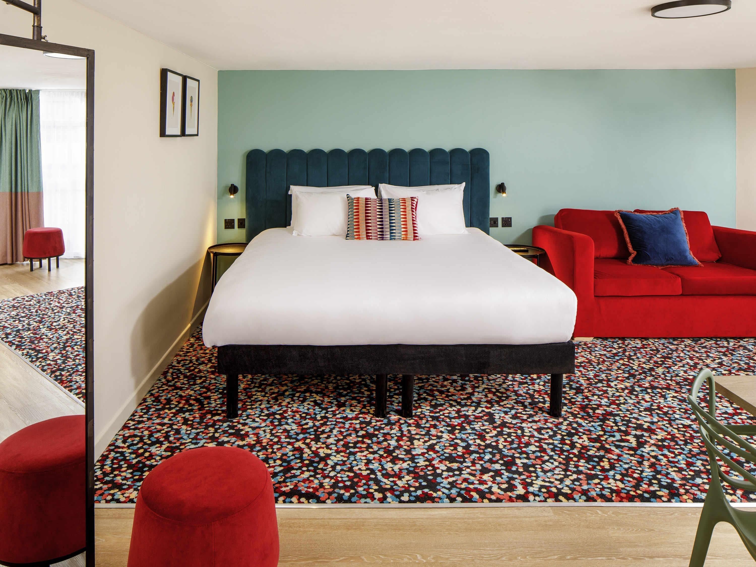 Une chambre d’hôtel avec un grand lit, un canapé rouge et une moquette aux motifs colorés.