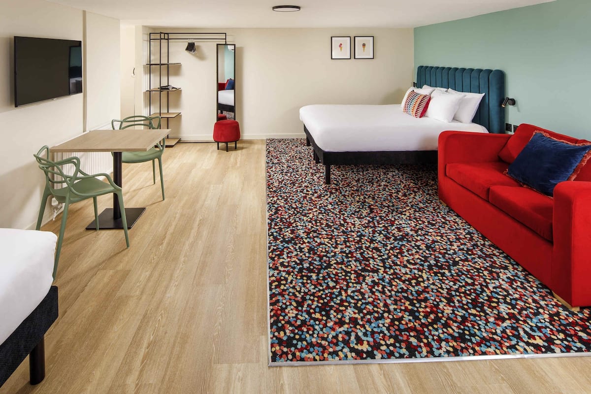 Quarto de hotel com uma cama, um sofá vermelho, uma mesa de jantar com cadeiras, uma televisão e um tapete estampado.