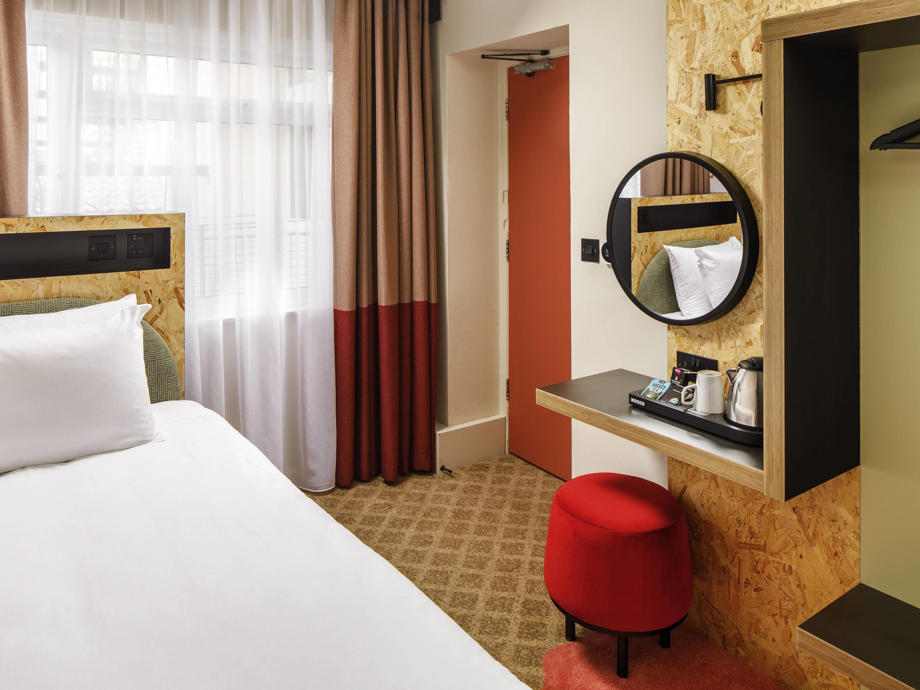 Une chambre d’hôtel avec un lit, un pouf rouge, une petite table avec une cafetière, un miroir et une fenêtre avec des rideaux légers.