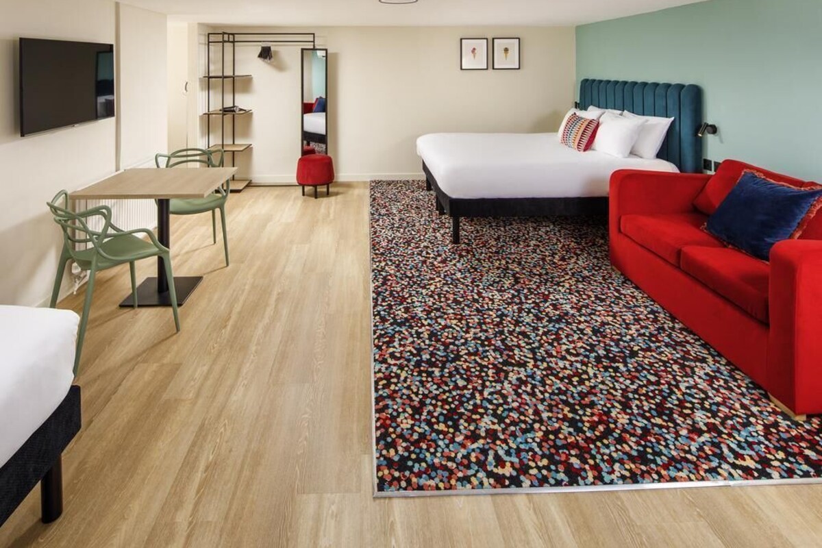 A hotel room with a bed, a red sofa, a small table, a TV, and a colorful rug.