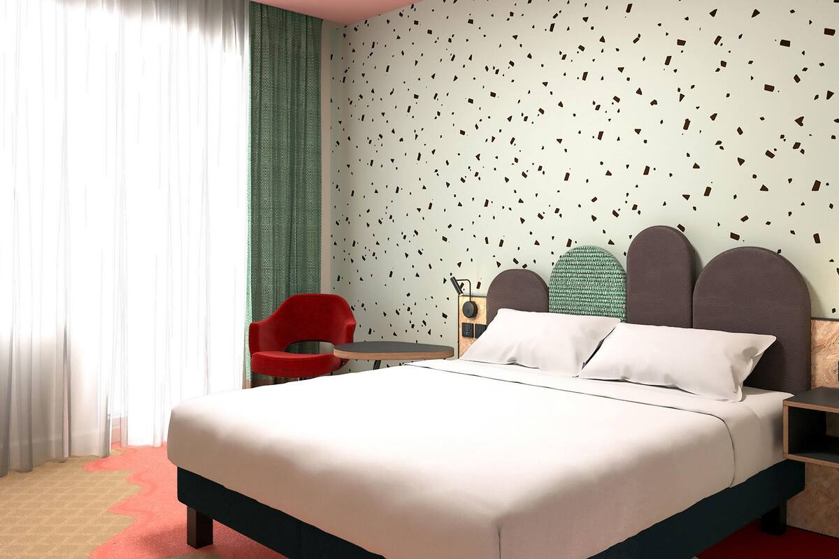 A modern hotel room with a bed, a red chair, a small table, and a wall with a geometric pattern.