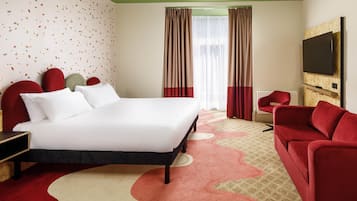 A hotel room with a bed, red upholstered headboard, a red sofa, a television, and a patterned carpet.