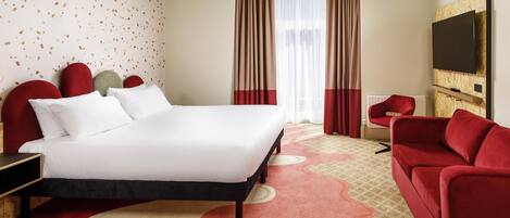 A hotel room with a bed, red upholstered headboard, a red sofa, a television, and a patterned carpet.