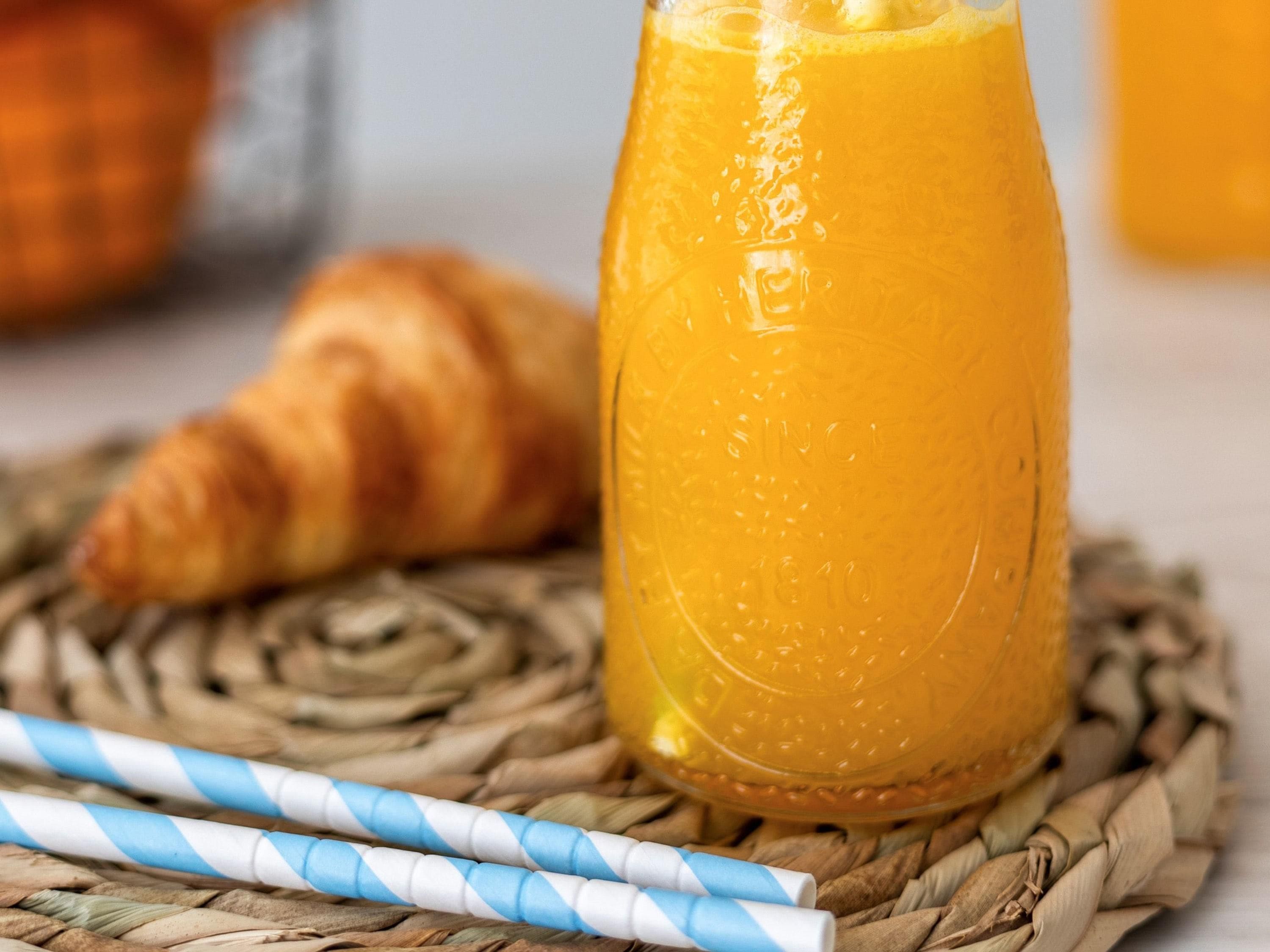 Een glas sinaasappelsap, een croissant en gestreepte rietjes op een geweven mat.