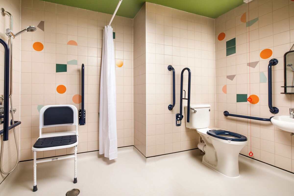 A bathroom with a wheelchair-accessible shower, a toilet with a safety bar, and a sink with a grab bar.
