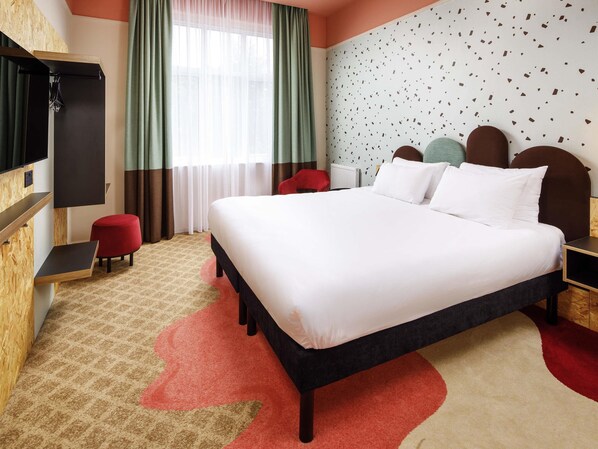 A hotel room with a large bed, a TV mounted on the wall, a red chair, and a window with curtains.