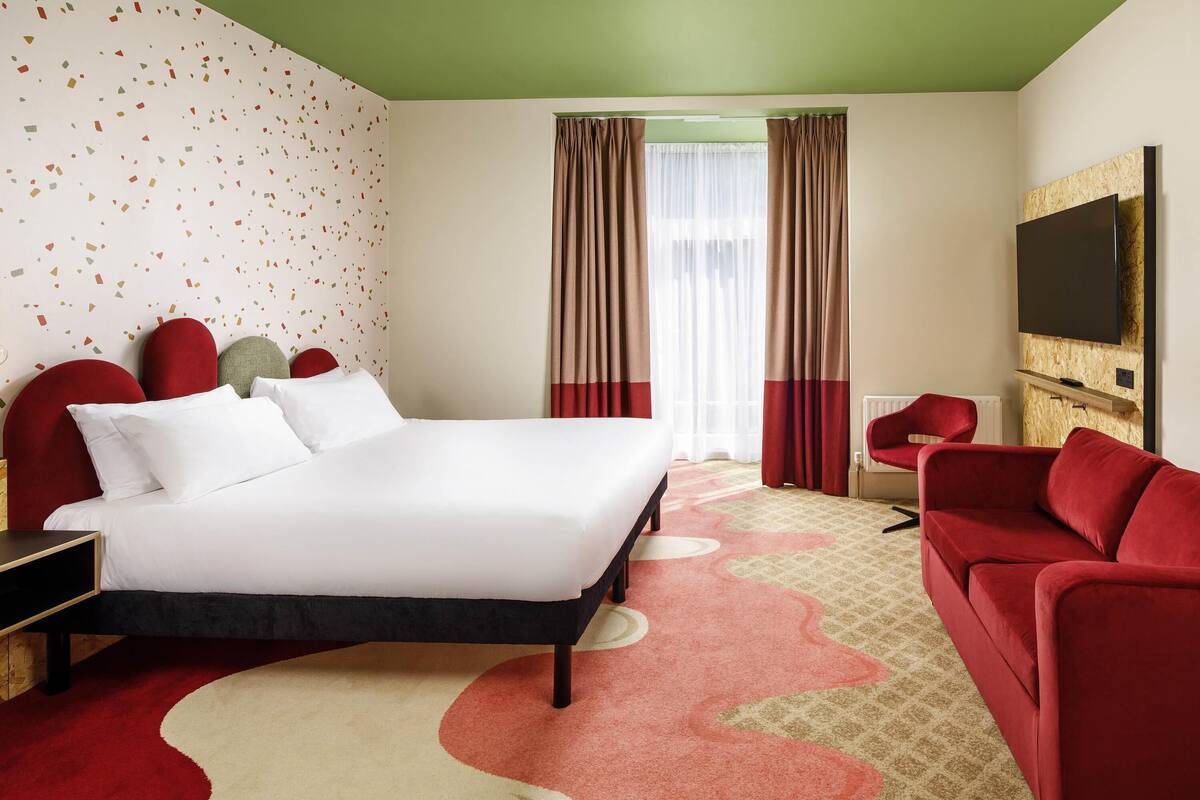 A hotel room with a bed, red upholstered headboard, a red sofa, a television, and a patterned carpet.
