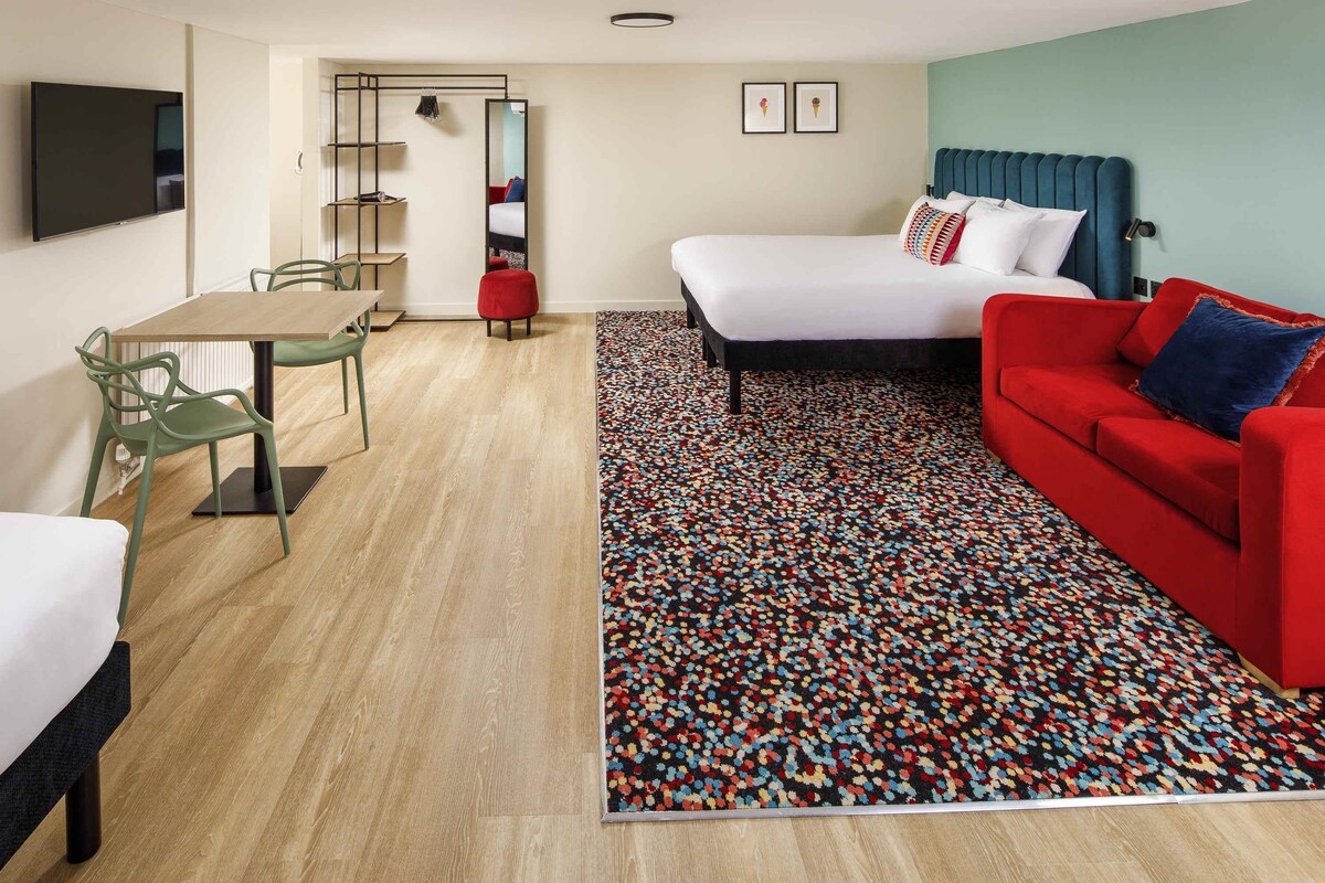 A hotel room with a bed, a red sofa, a dining table with chairs, a TV, and a patterned rug.