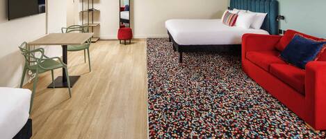 Habitación de hotel con cama, sofá rojo, mesa de comedor con sillas, televisión y alfombra estampada.