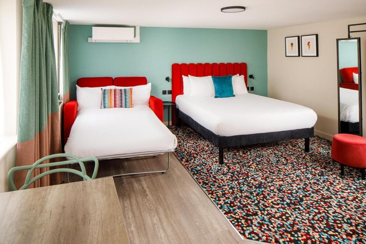A hotel room with two beds, a small table, a chair, a mirror, and a colorful carpet.