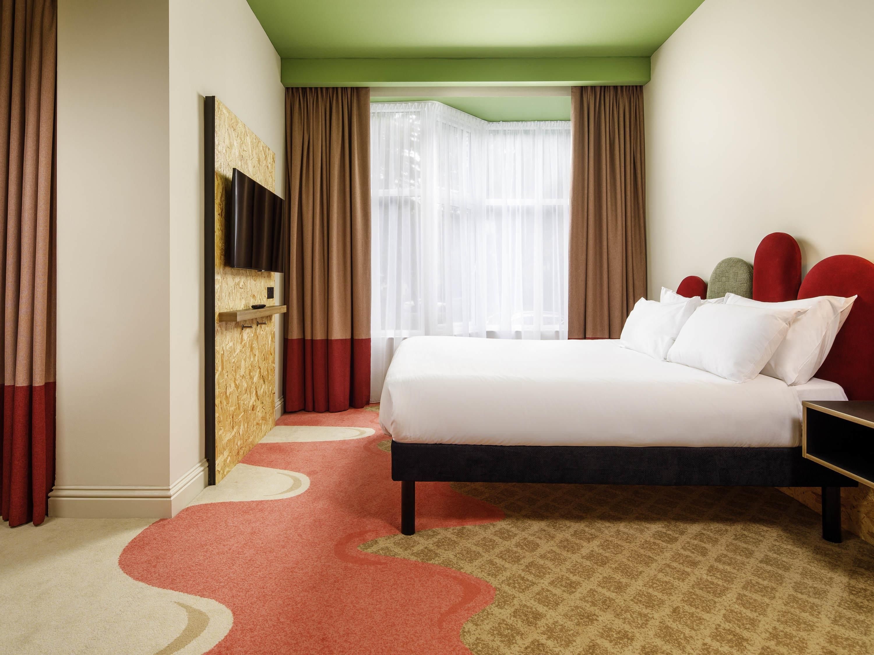 Une chambre d’hôtel avec un lit, un téléviseur fixé au mur et une fenêtre avec des rideaux.