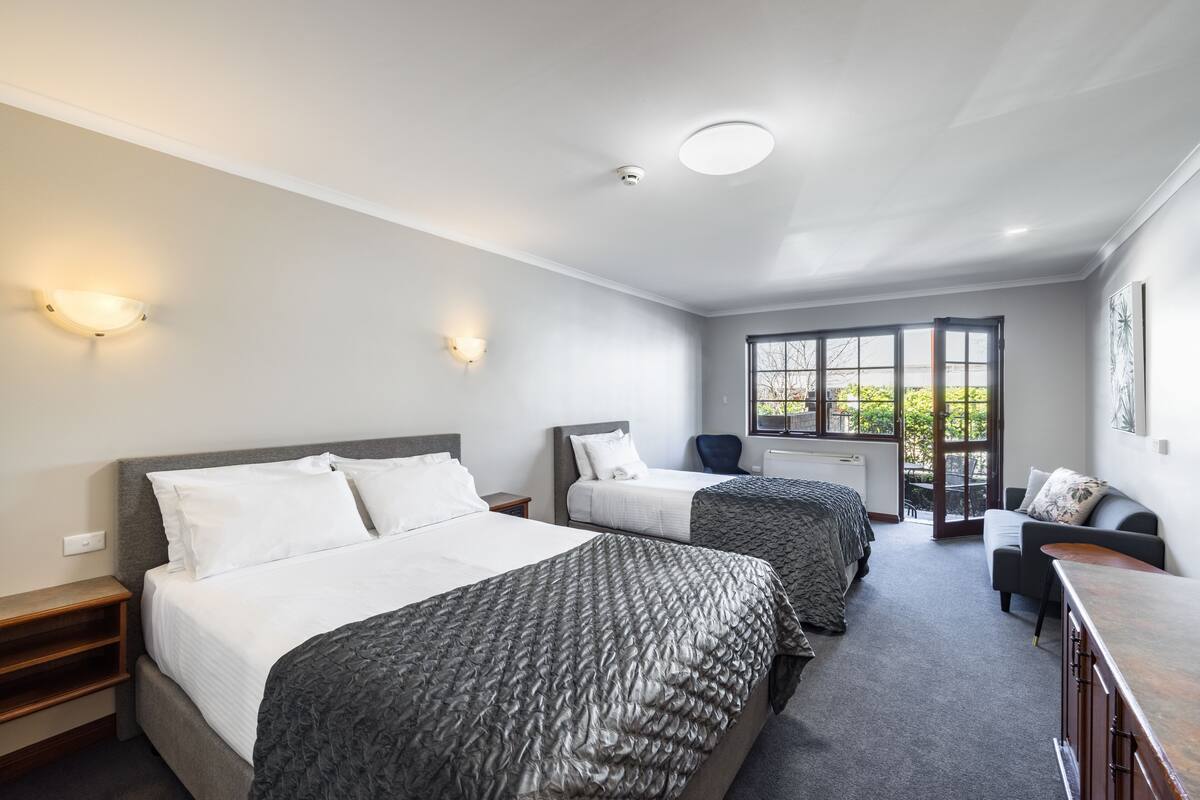 A modern hotel room with two beds, a sofa, a desk, and a large window.