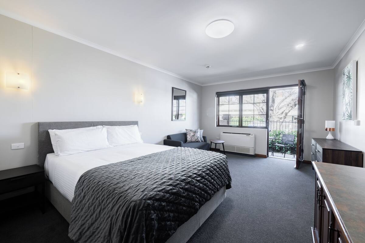 A modern bedroom with a large bed, bedside tables, a small seating area, and a view of the outdoors.