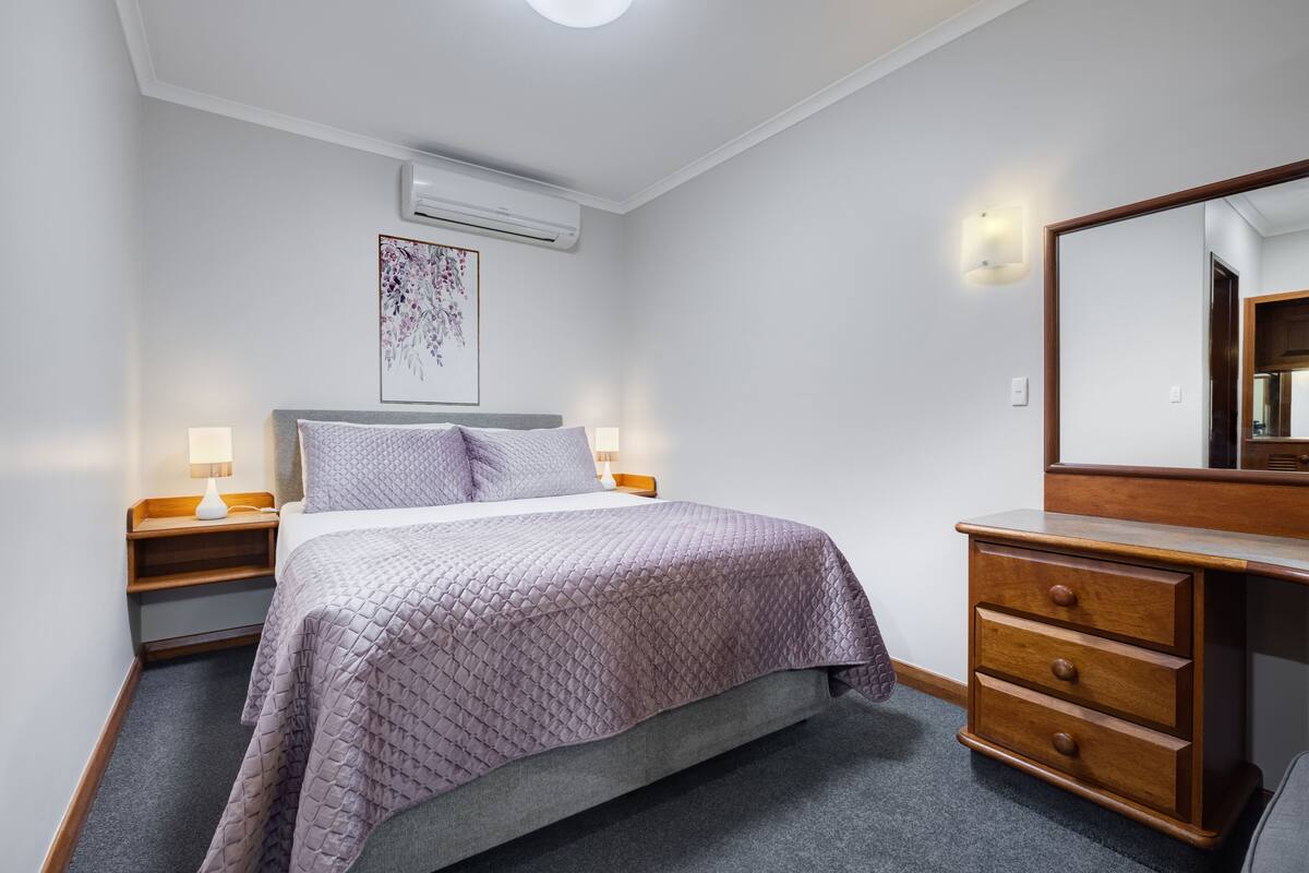 A bedroom with a bed, bedside tables, a dresser, a mirror, and an air conditioning unit.
