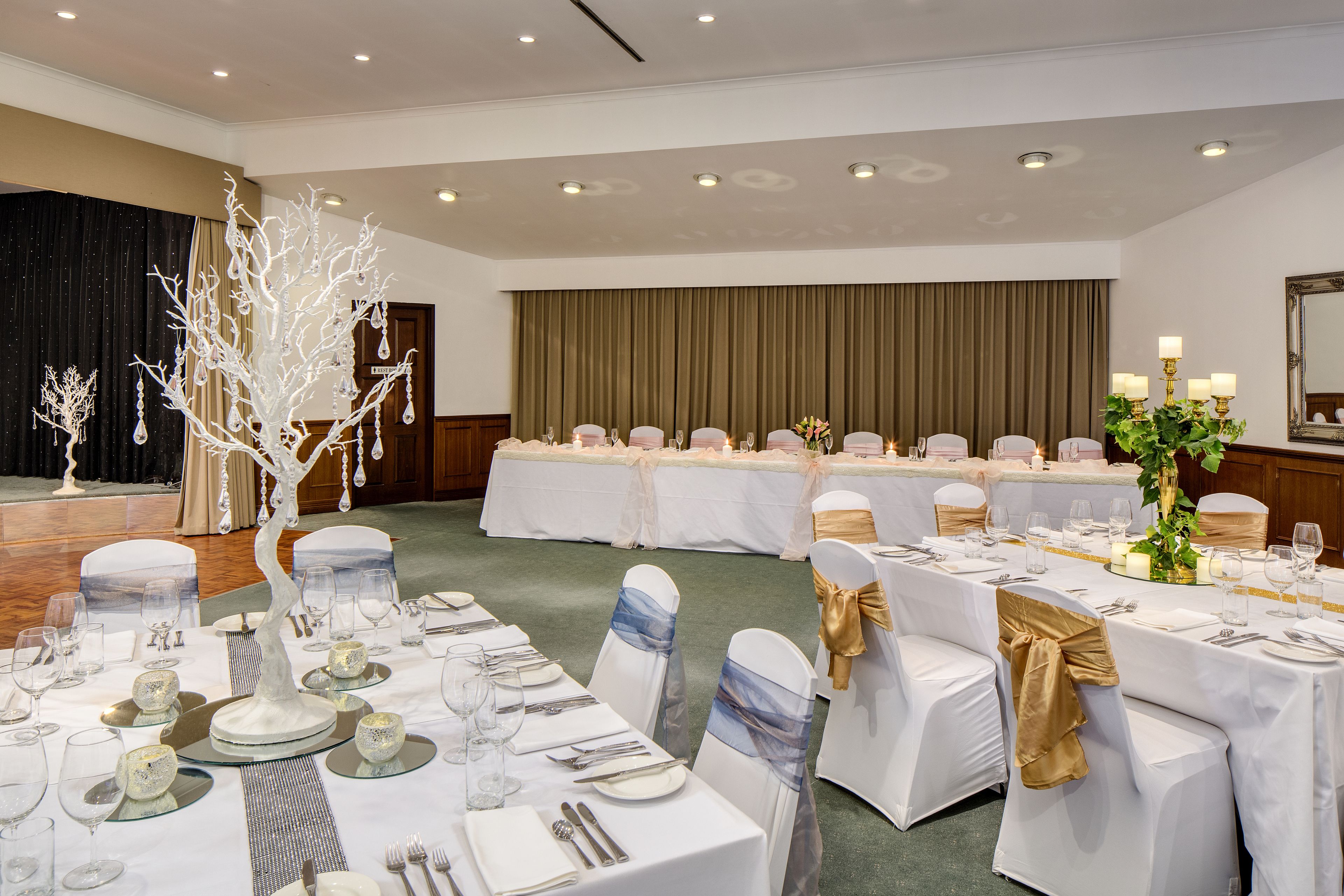 Un salón de banquetes con mesas redondas dispuestas para un evento formal, con manteles blancos, cintas doradas y un centro de mesa decorativo con un árbol blanco.