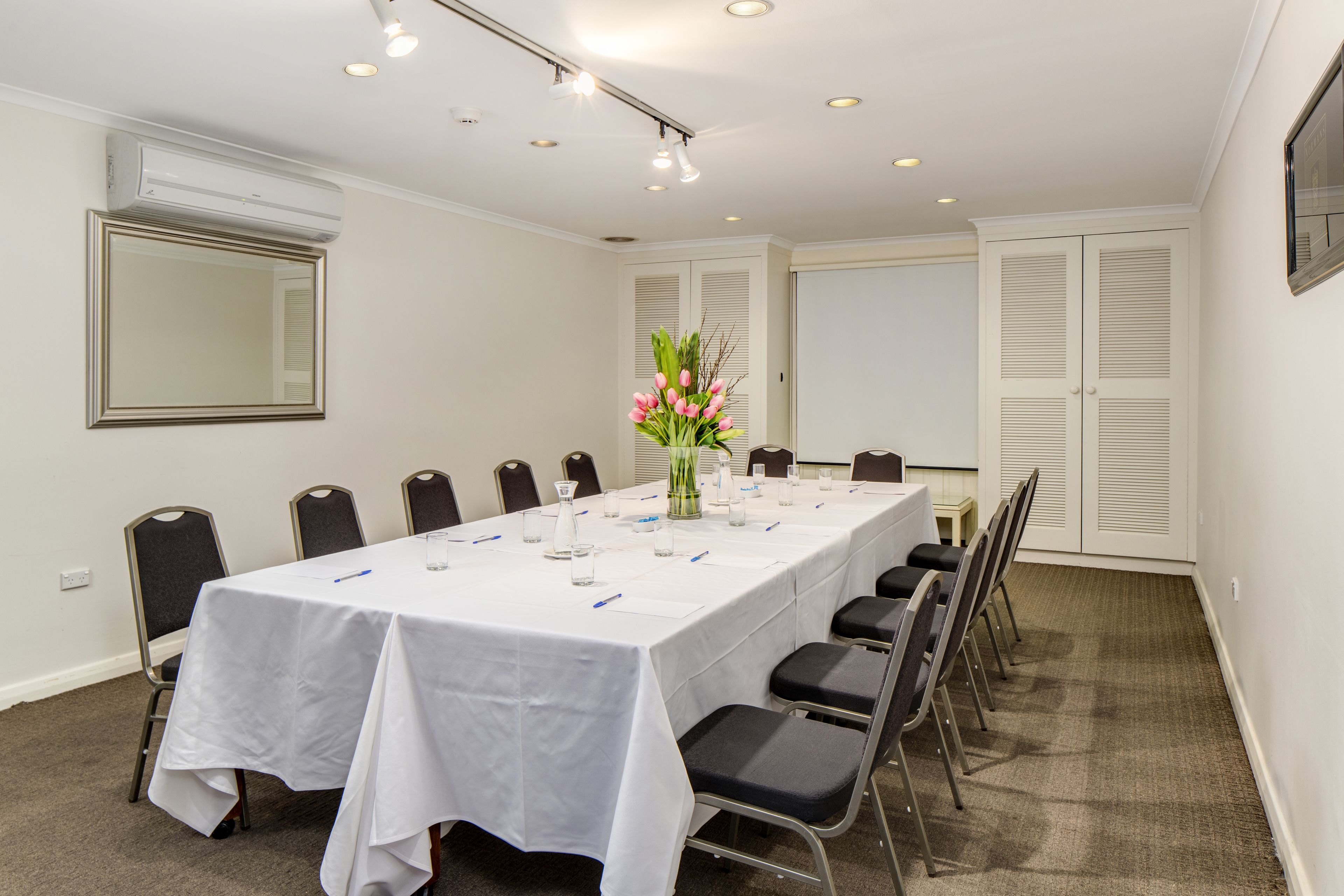 Une salle de conférence avec une longue table, des chaises, un vase de fleurs et un grand miroir au mur.