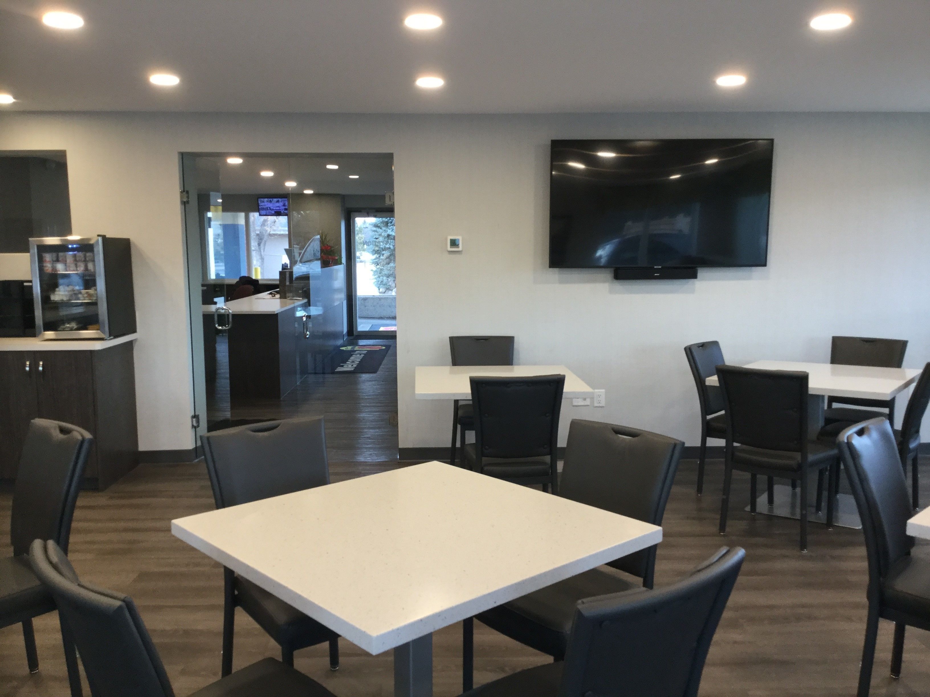 A modern dining area with white tables and black chairs, a flat-screen TV mounted on the wall, and a refrigerator in the background.