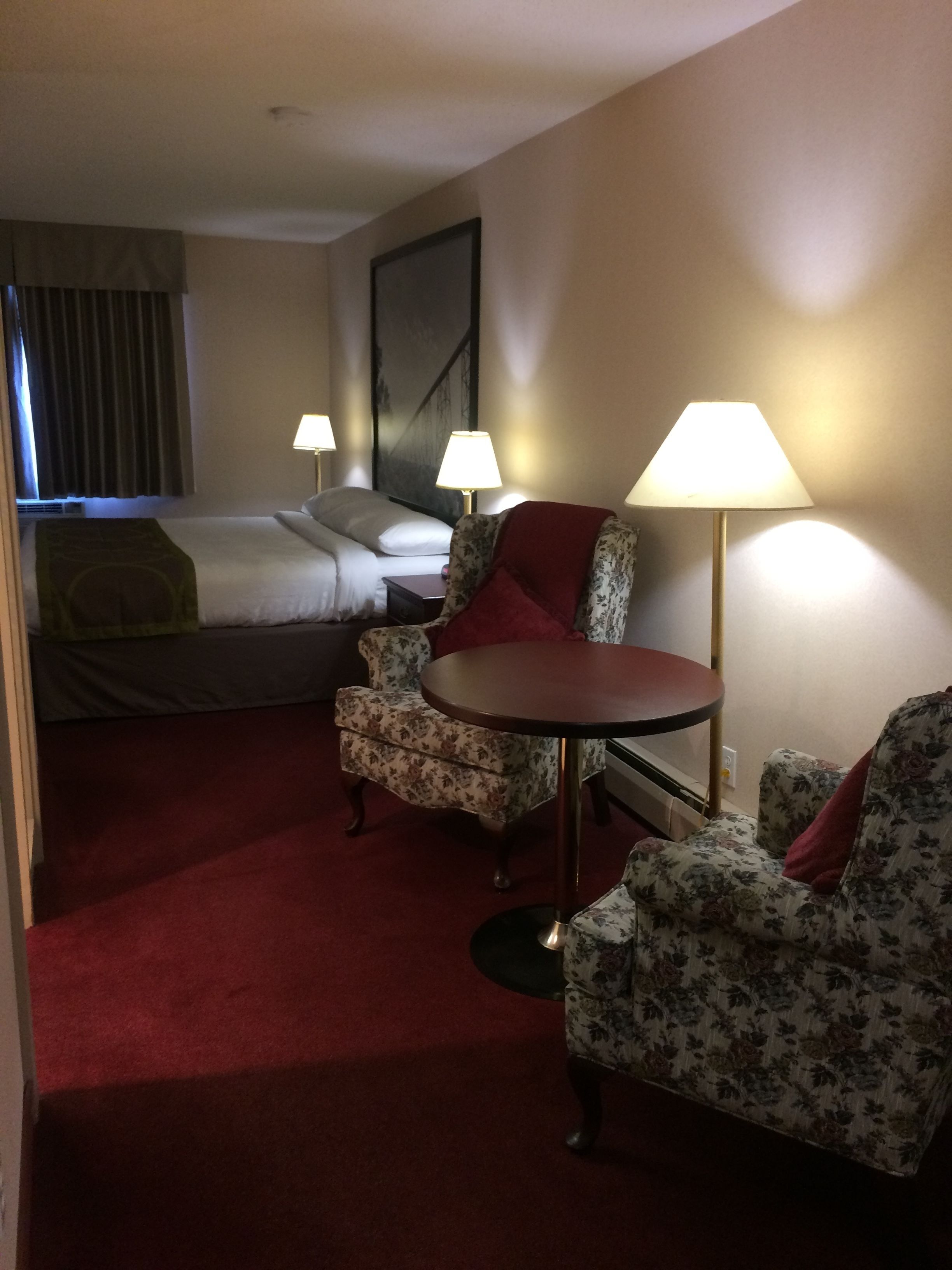 A hotel room with a bed, two chairs, a small table, and two lamps.