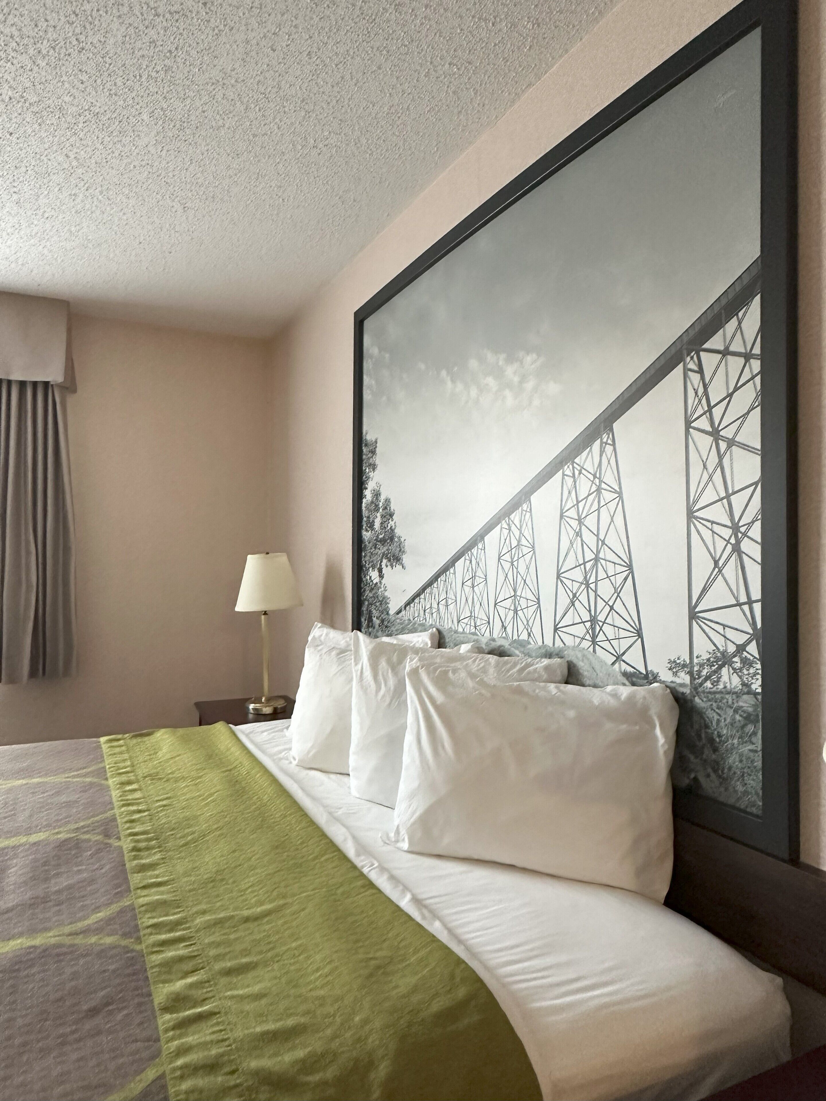Quarto de hotel com uma cama, mesa de cabeceira, abajur e uma grande moldura com uma fotografia de uma ponte.