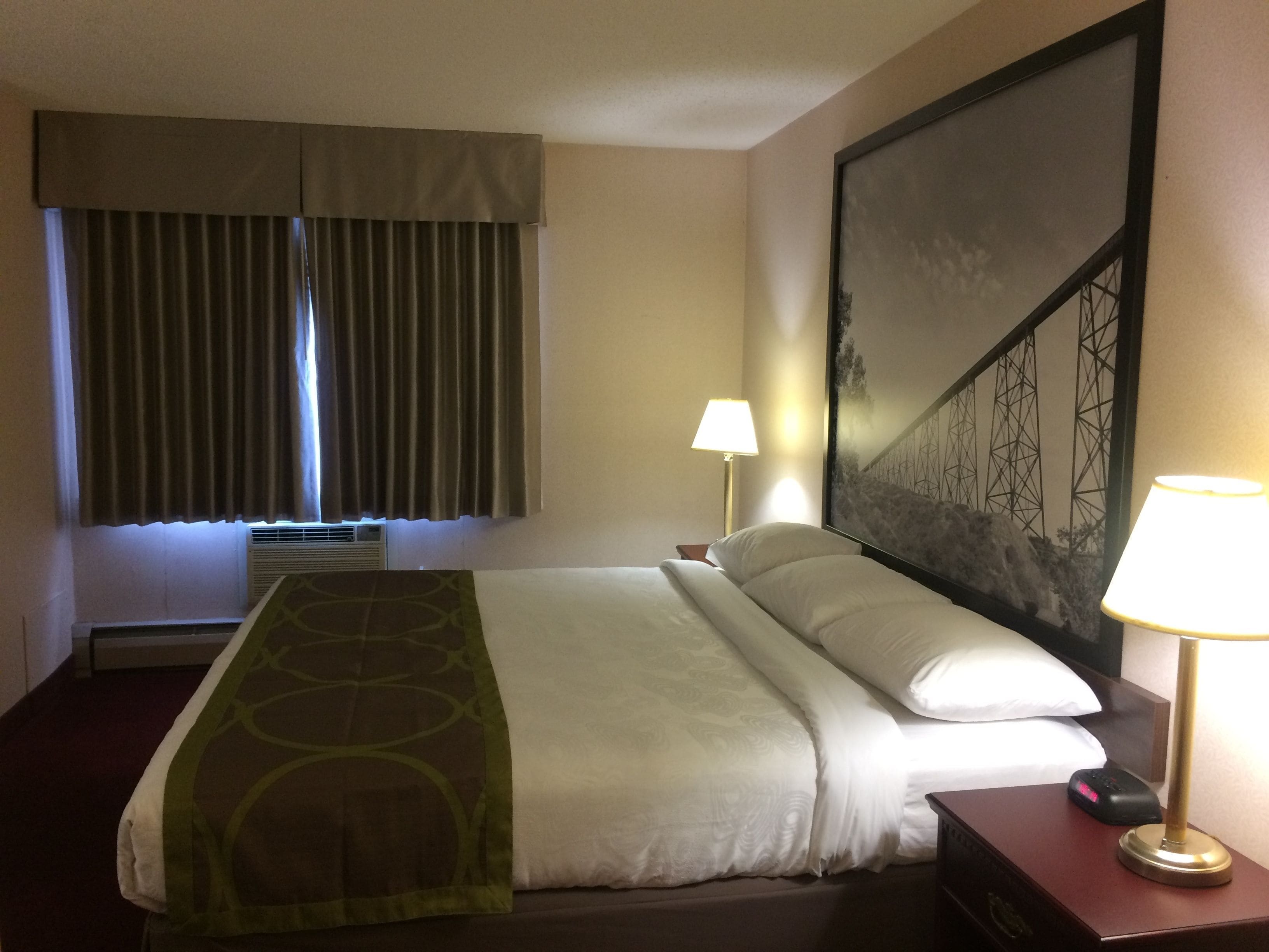 A hotel room with a bed, two bedside lamps, a large painting on the wall, and a window with curtains.