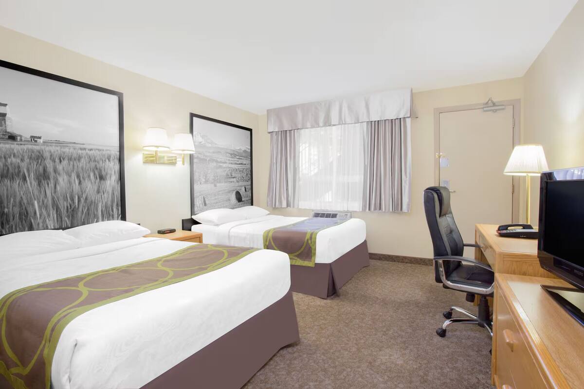 Habitación de hotel con dos camas, un escritorio con televisor, una silla y un ventanal con cortinas.