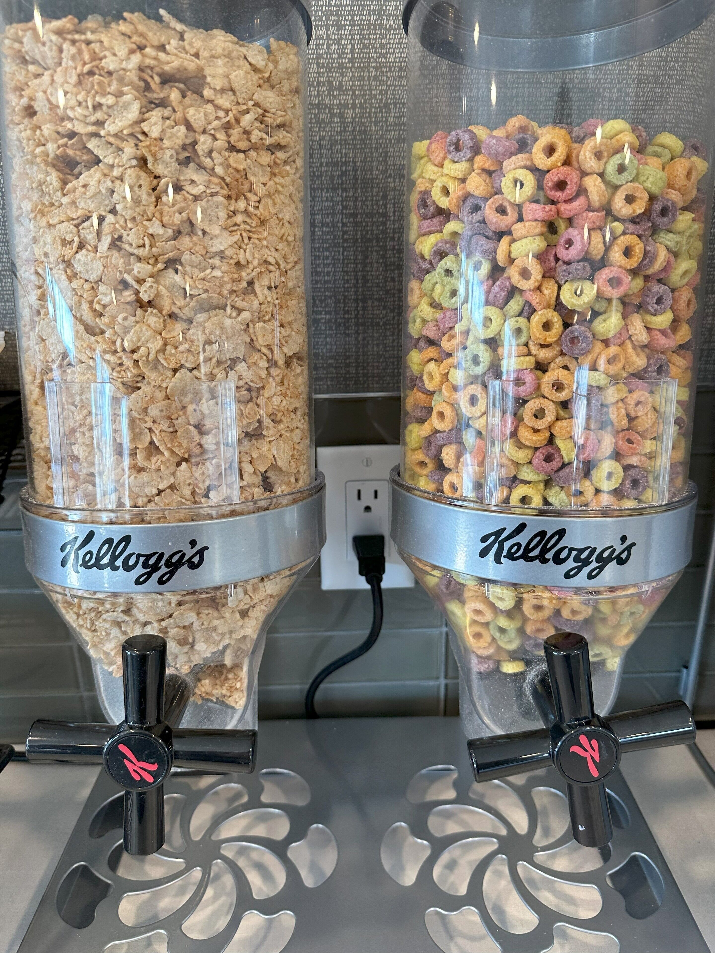 Two clear plastic containers labeled 'Kellogg's' filled with cereal flakes and colorful cereal rings, placed on a metal surface with a power outlet in the background.
