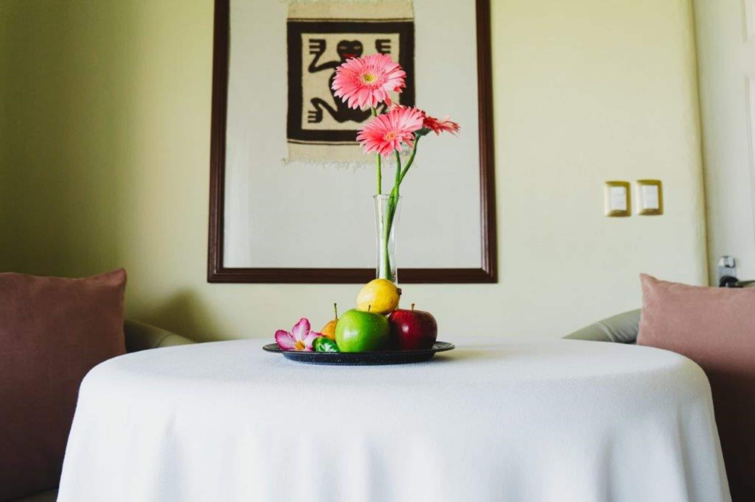 Ein Tisch mit einer Vase voller rosa Blumen und Früchten, ein gerahmtes Bild an der Wand und eine weiße Tischdecke.