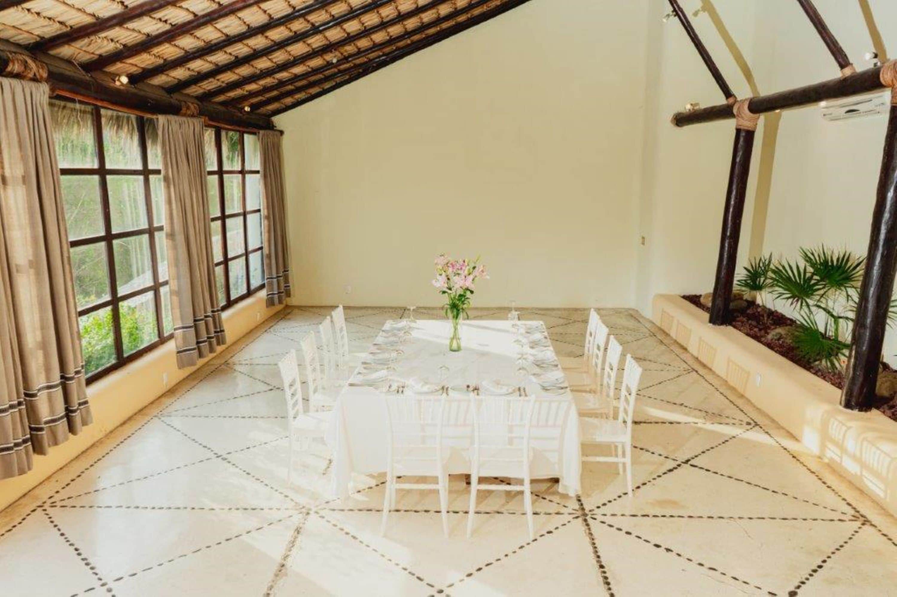 Una habitación amplia con techo alto, una gran mesa de comedor preparada para seis personas y vistas a zonas verdes a través de amplios ventanales.