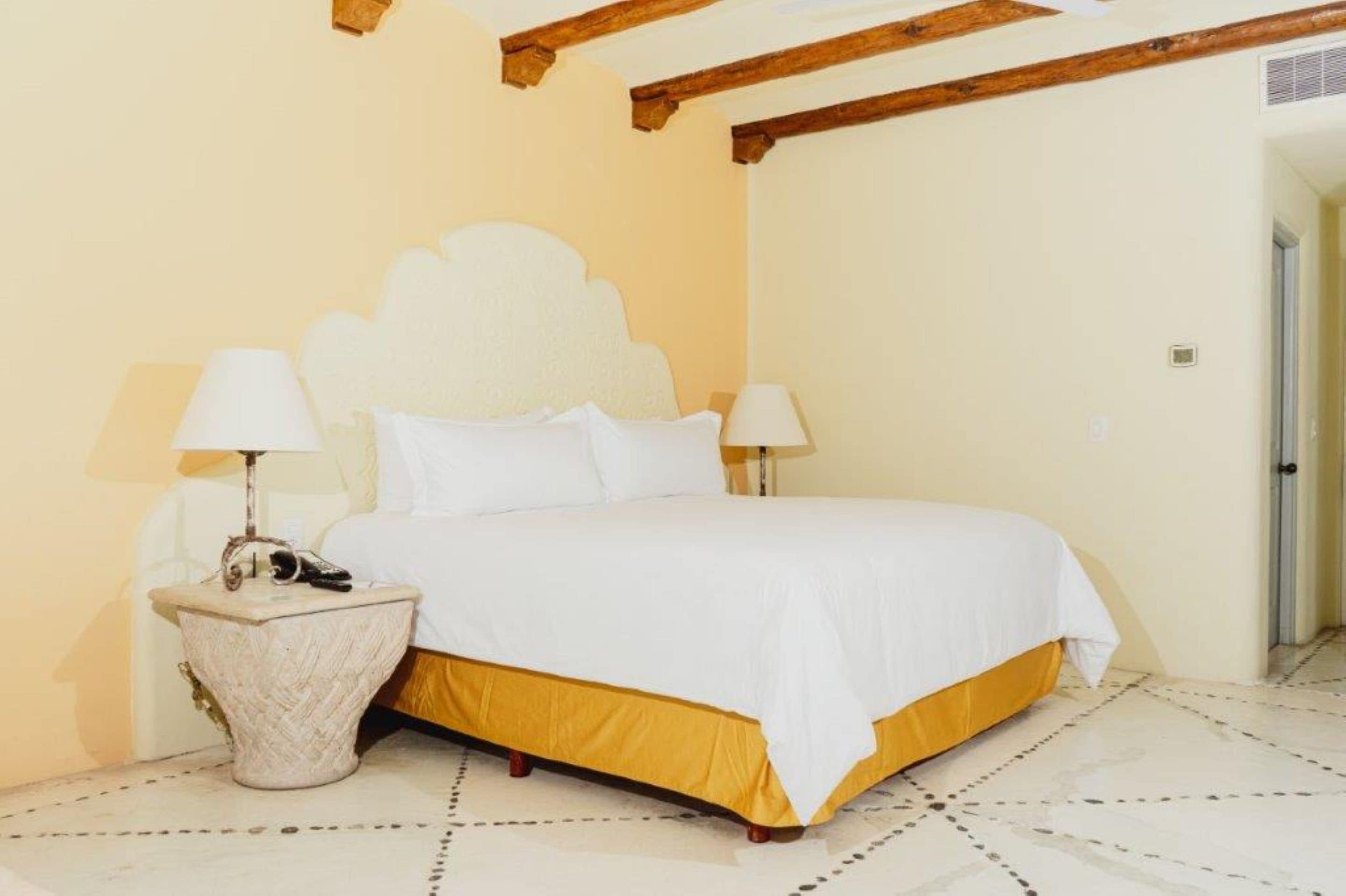 Quarto com cama, duas luminárias de cabeceira, uma mesinha de apoio e um vigário de madeira no teto.