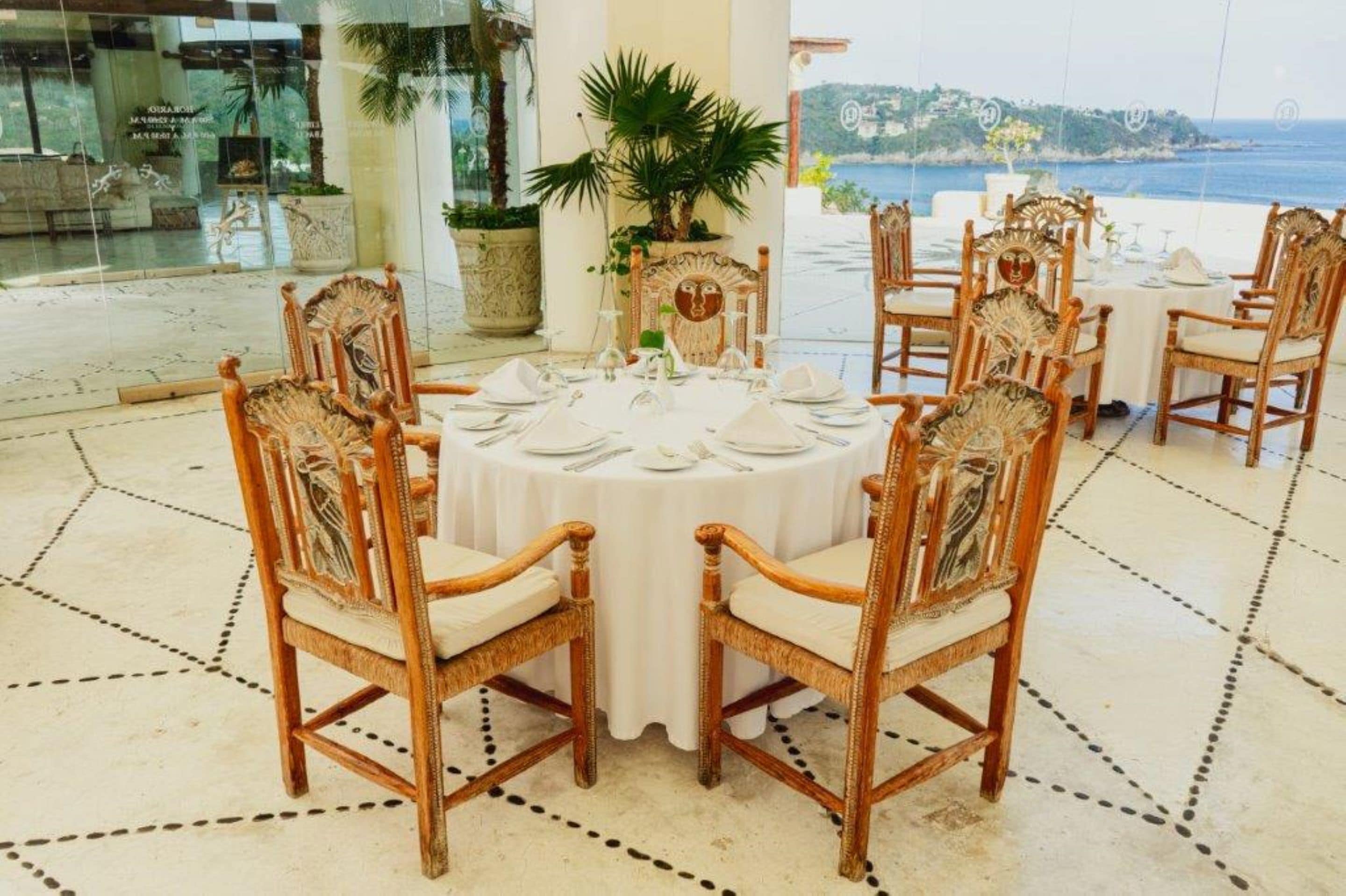 Une table ronde dressée pour six personnes, agrémentée de chaises décorées, d’une plante en pot et offrant une vue sur la mer.