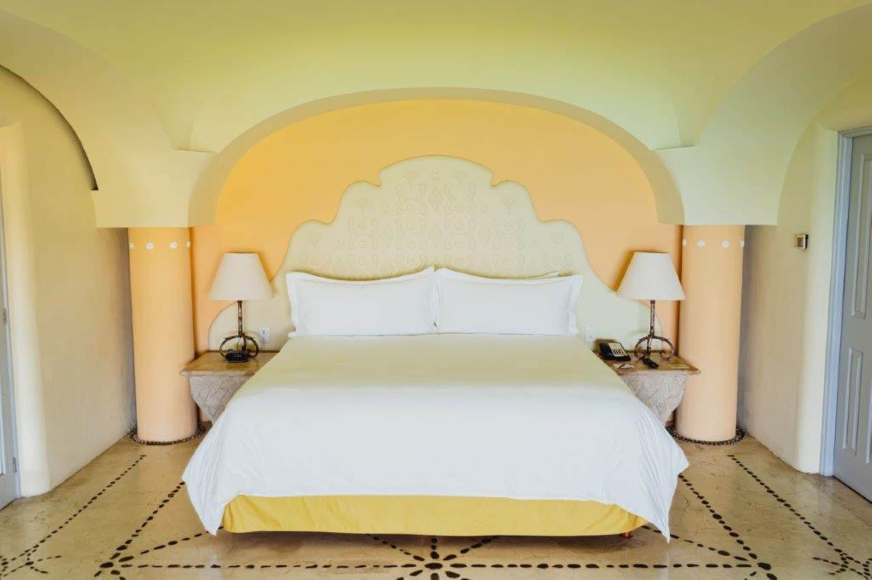 Una cama grande con cabecera blanca, dos lámparas de noche y un suelo estampado.