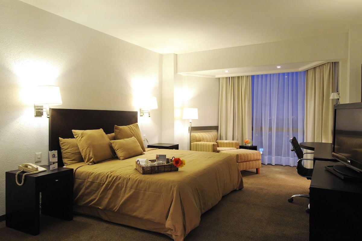 A hotel room with a large bed, bedside tables, a desk with a chair, a TV, and a window with curtains.