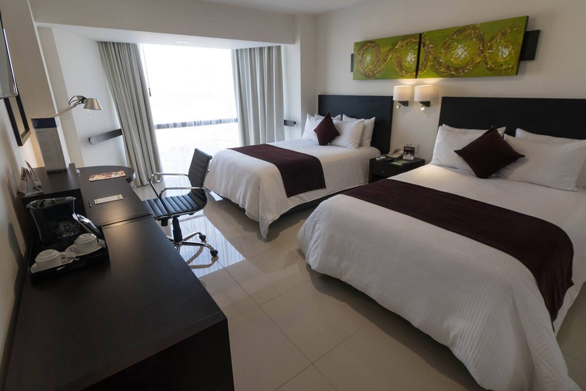 A hotel room with two beds, a desk with a coffee maker, a chair, and a view of the outdoors.