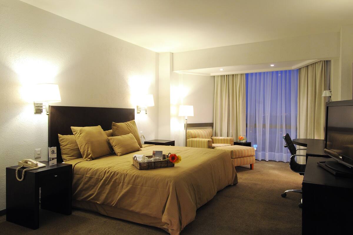 A hotel room with a large bed, two bedside tables, a desk with a chair, a television, and a window with curtains.