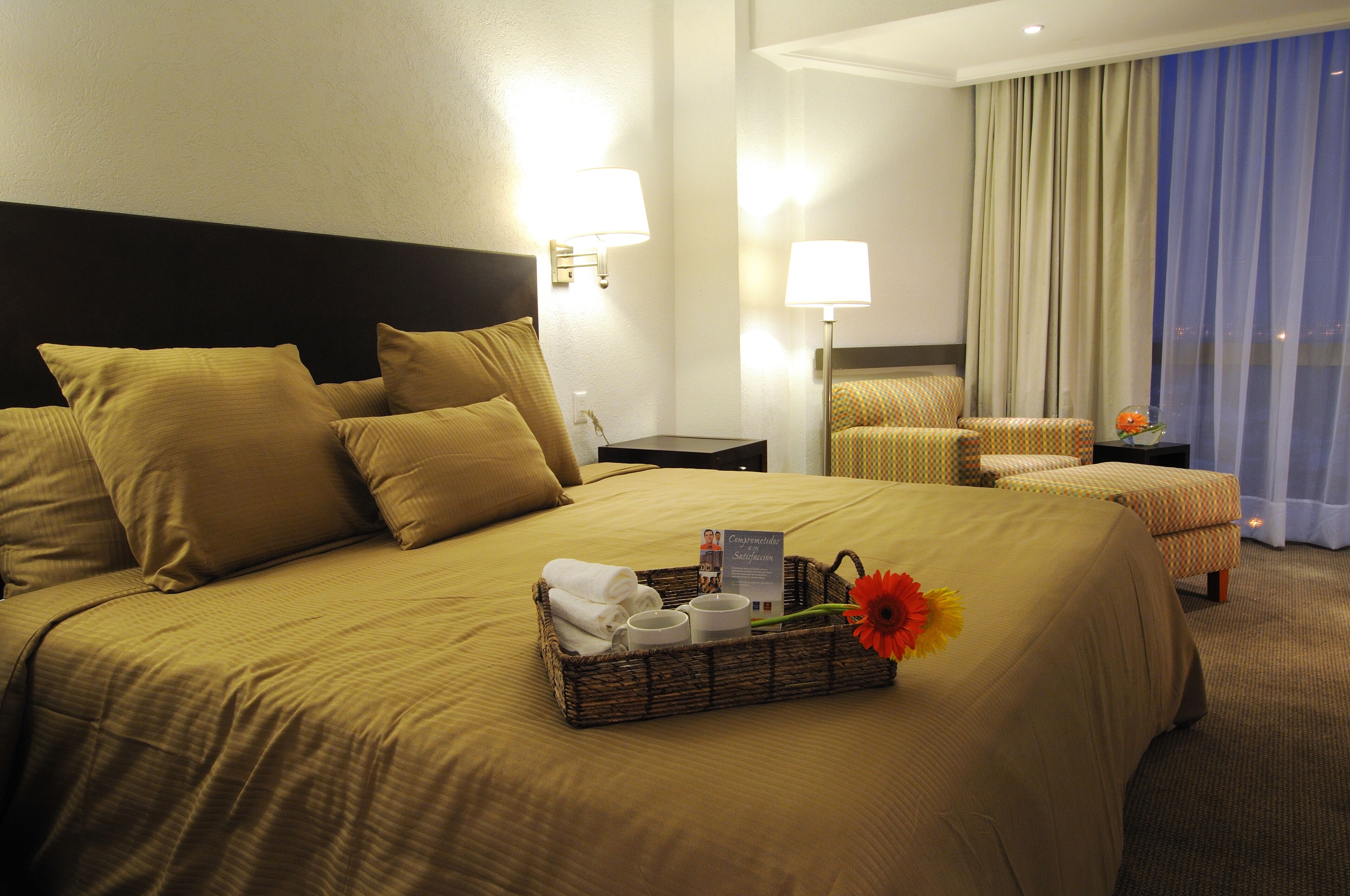 A hotel room with a large bed, two bedside lamps, a chair, a small table, and a window with curtains.