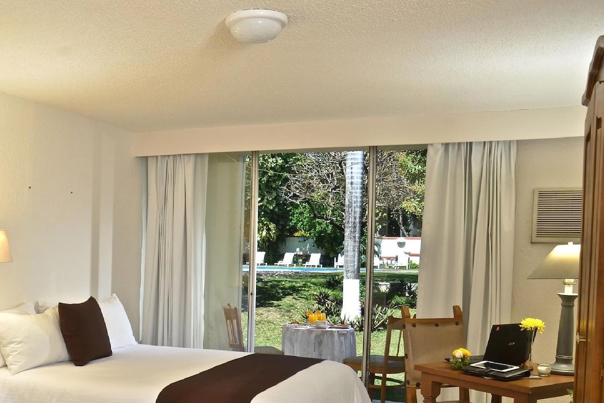 Una habitación de hotel con cama, escritorio con ordenador portátil, silla y ventana con cortinas.