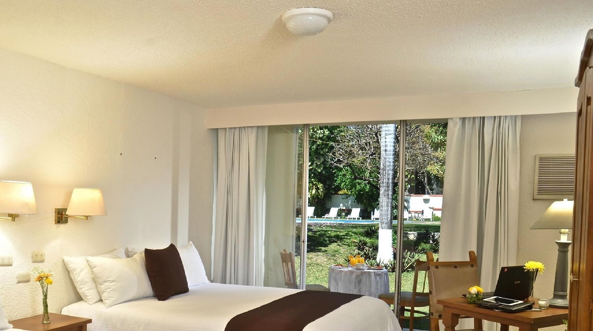 Een hotelkamer met een bed, een bureau met een laptop, een stoel en een raam met gordijnen.