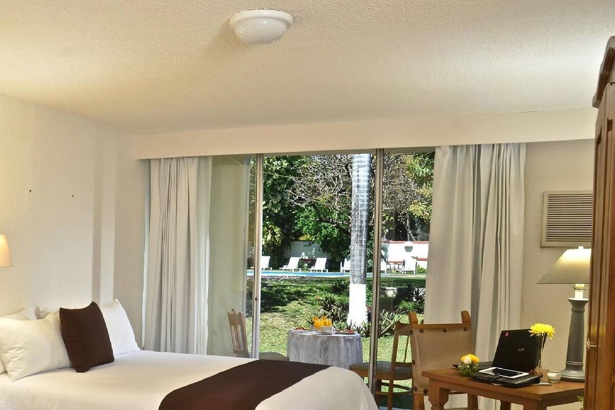 Quarto de hotel com cama, secretária com laptop, cadeira e janela com cortinas.